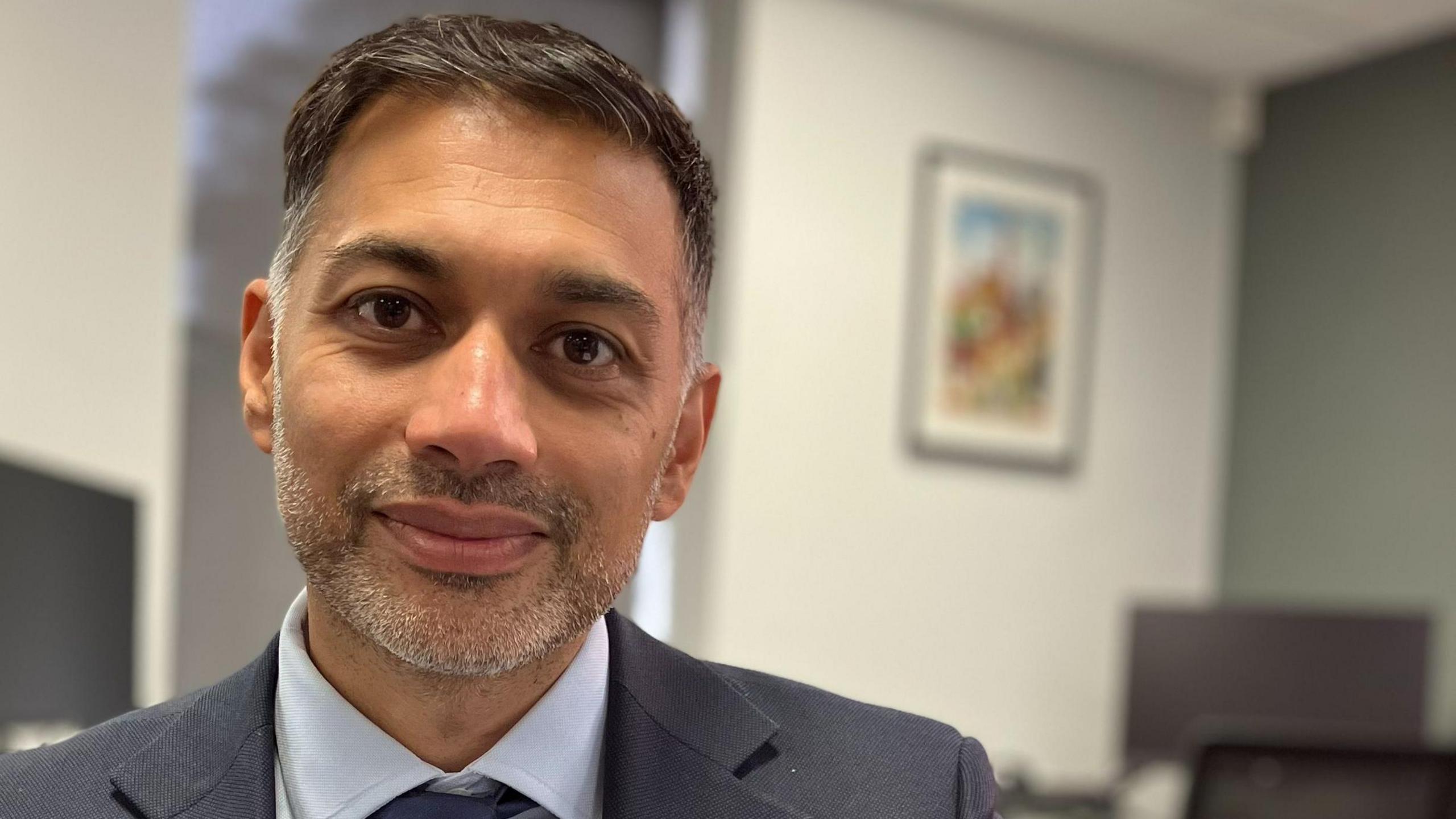 Arun Chauhan sits in an office with white and green walls. He has short dark hair, a short grey beard and wears a grey suit, light-grey shirt and blue tie.