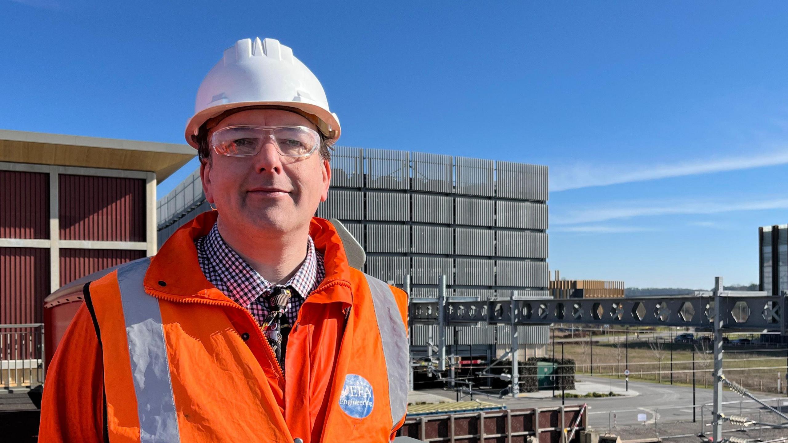 Philip Holbourn is standing on the construction site wearing a white hard hat, safety glasses and an orange high viability coat over a shirt and tie. Its is a sunny day with blue skies. 