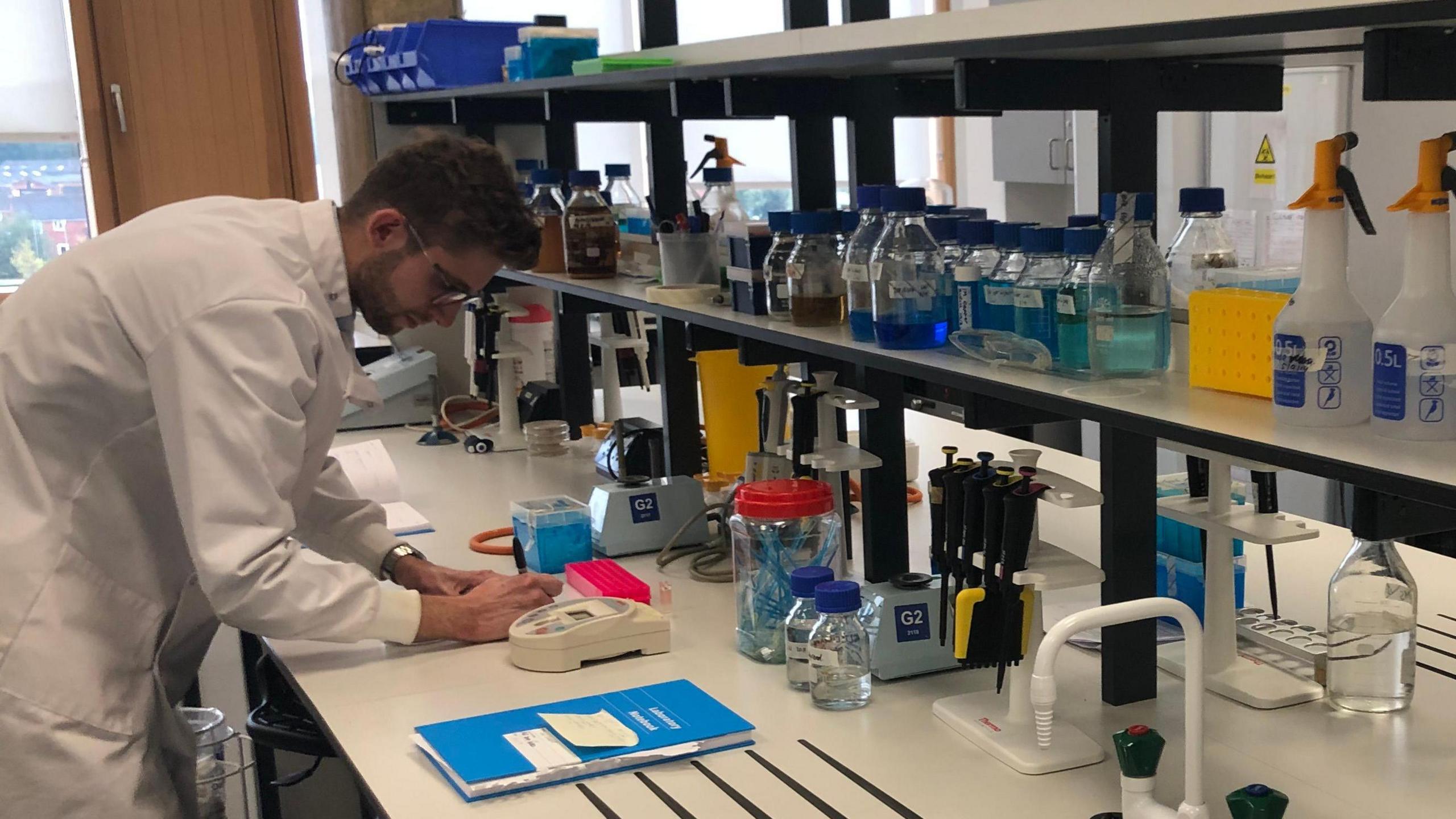 A man in a white lab coat and glasses writes on a table with lots of lab equipment on a large white table in front of him. 