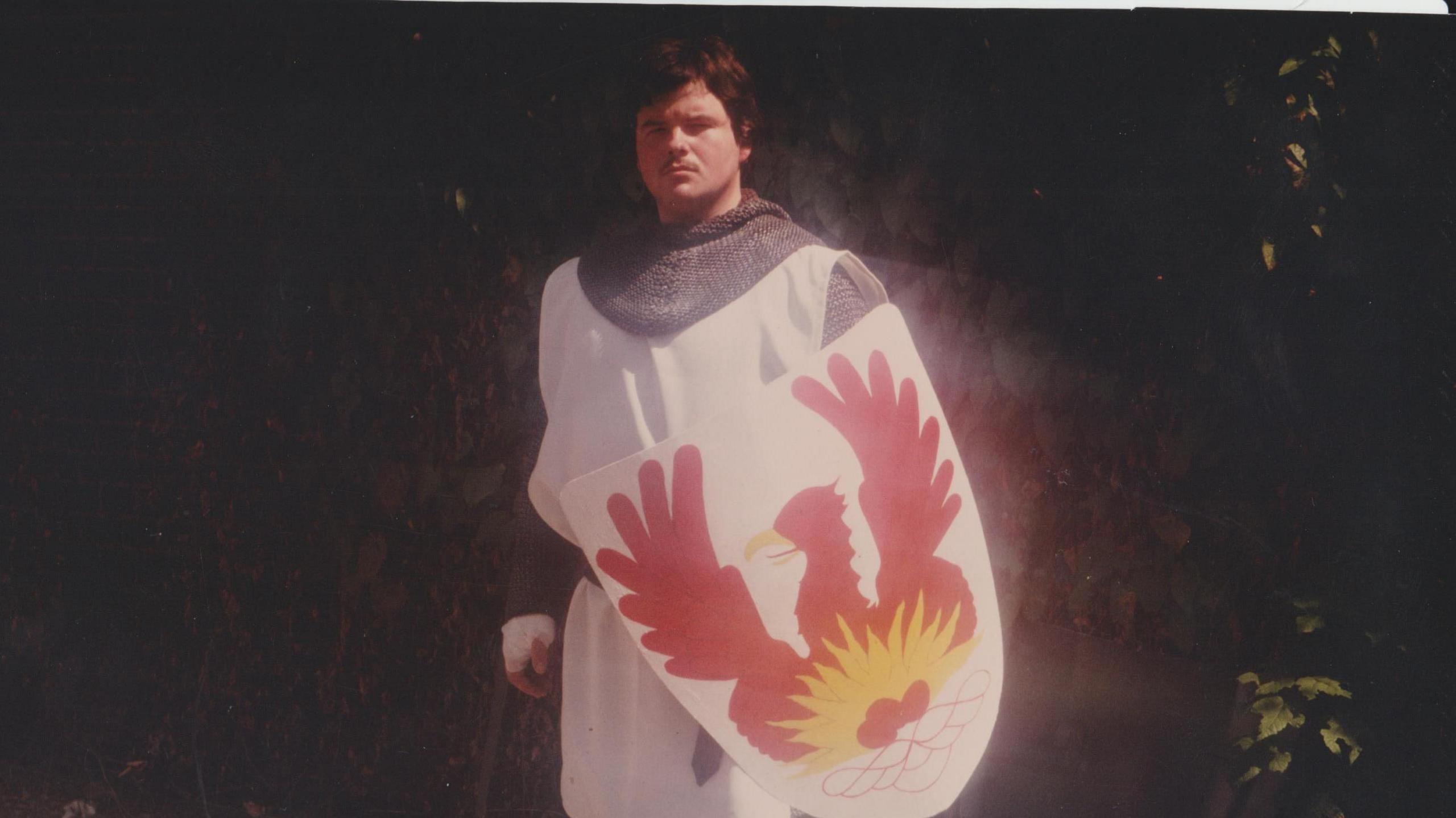 A man with brown hair dressed as a knight with a shield which has a red eagle with its wings spread on it.