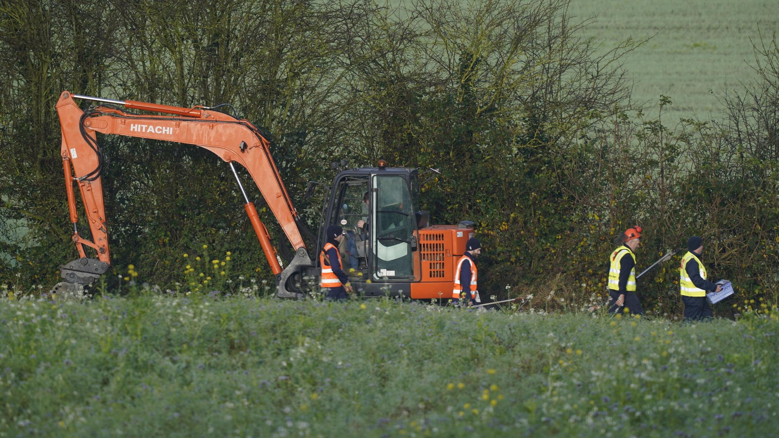 A field with trees in the background. In front of the trees is an orange digger and four workmen. 