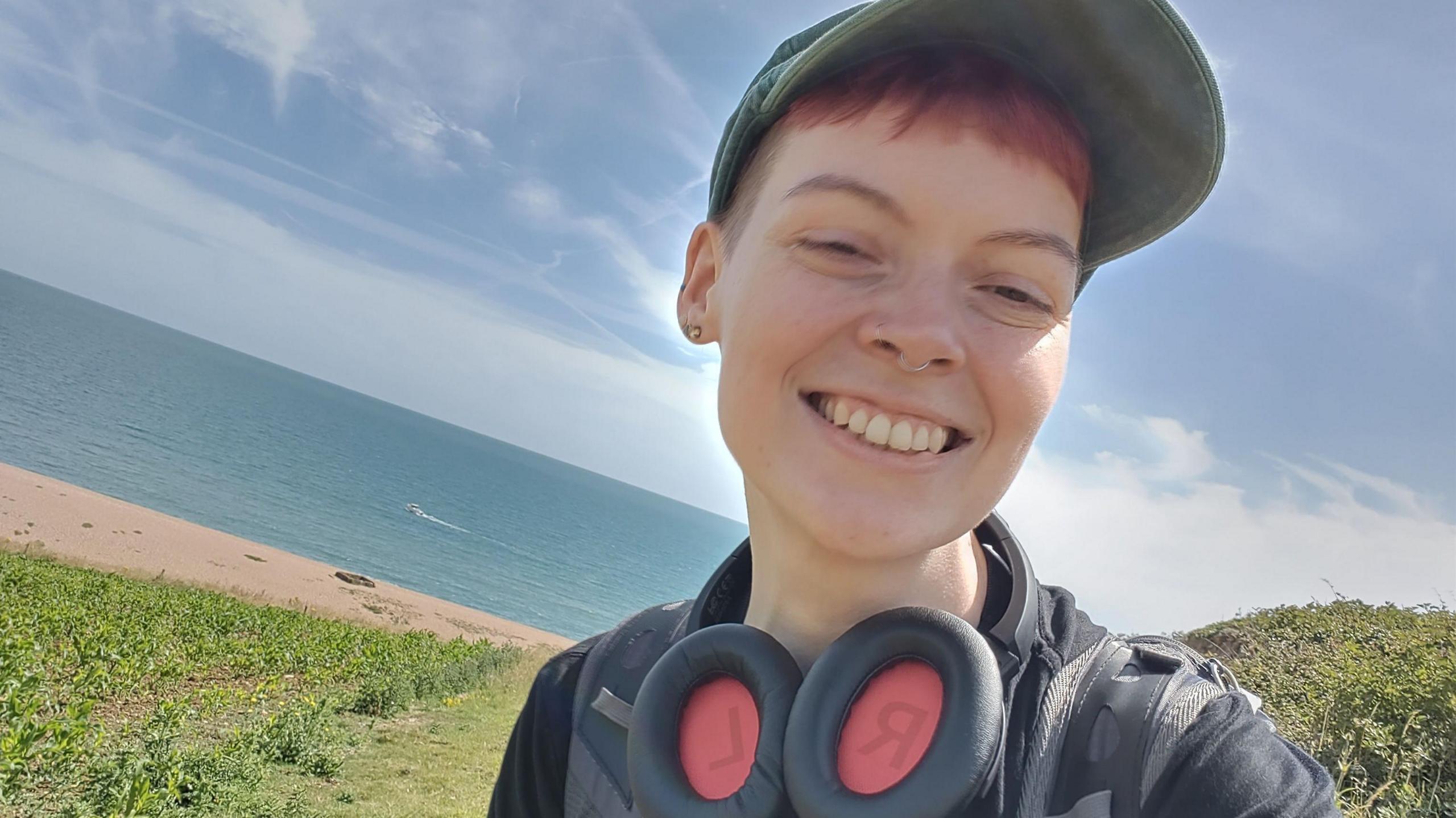 A woman stood on a hill with a beach and the sea behind her. She is smiling and has on a baseball cap and large headphones around her neck. 