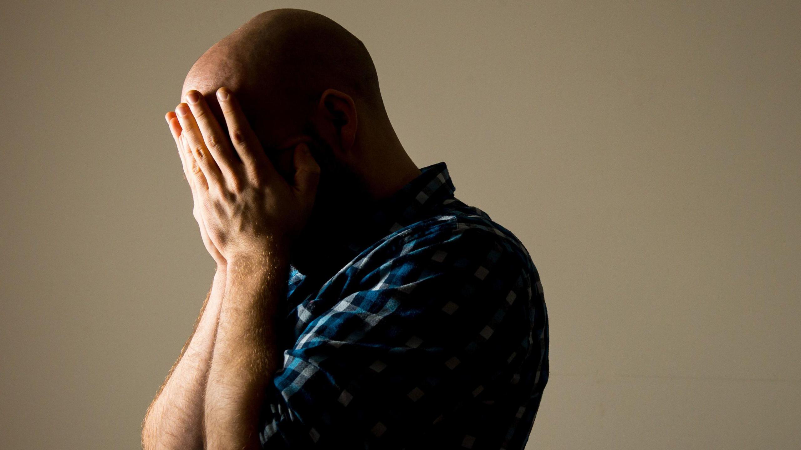 A bald man wearing a blue chequered shirt with his head in his hands.