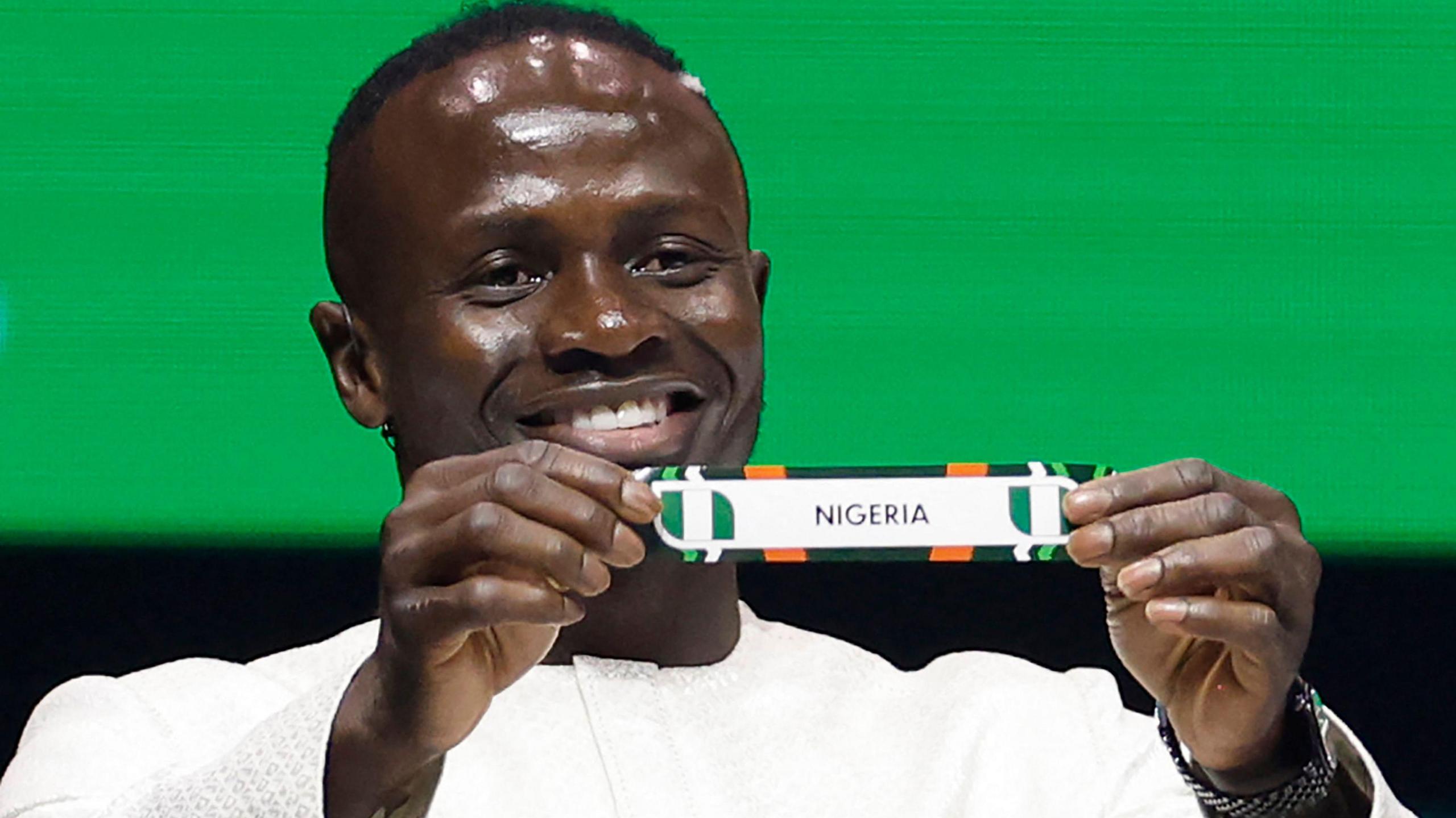 Sadio Mane, wearing a white tunic and standing in front of a green and black background, smiles as he holds a rectangular piece of paper bearing the word Nigeria and the Nigerian flag on it