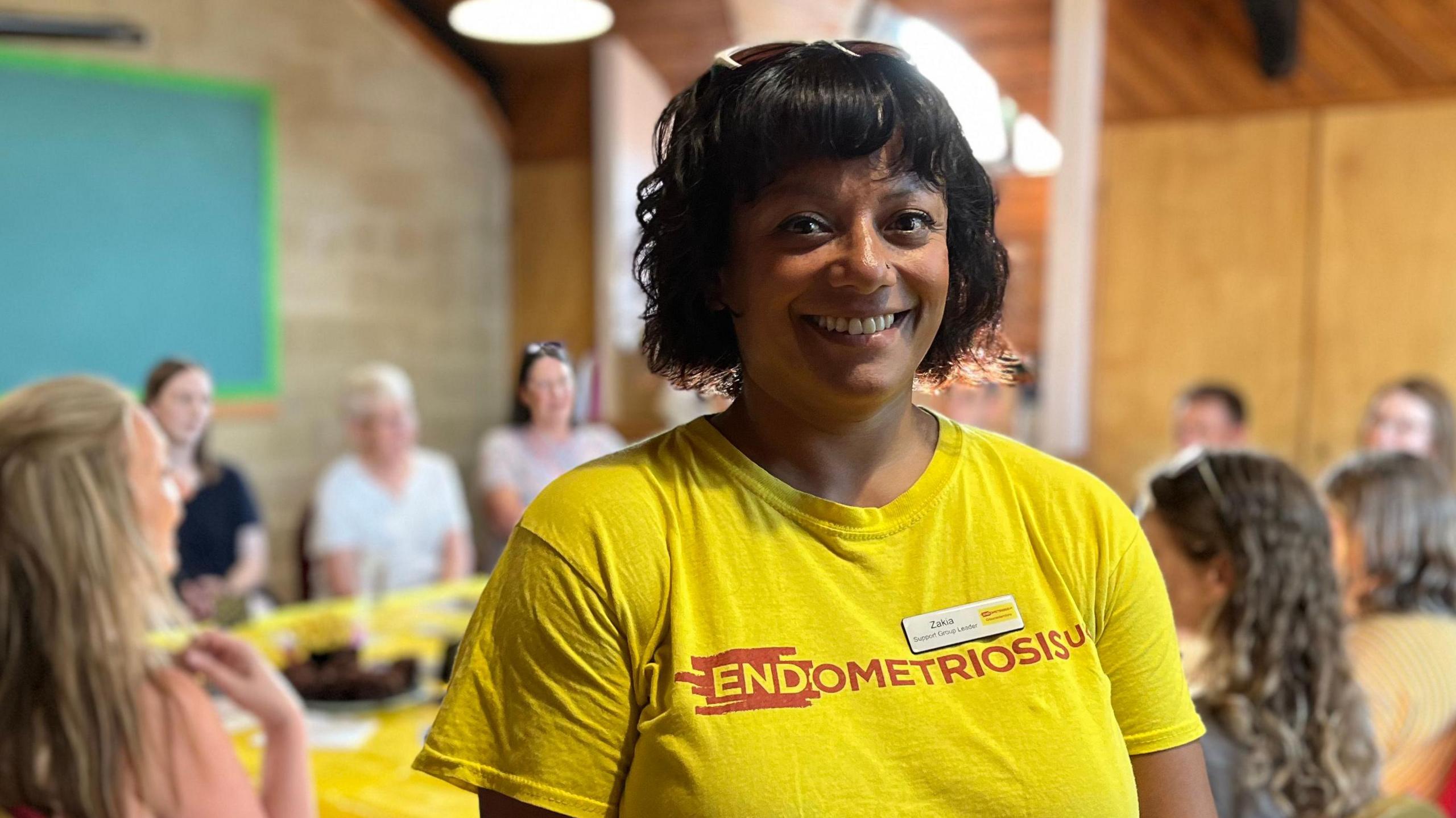 Jakia smiles at the camera, wearing a bright yellow top with the words "Endometriosis UK" on it in red writing. She has a dark brown bob with a fringe, and wears sunglasses on top of her head. Behind her, the support group can be seen talking. 
