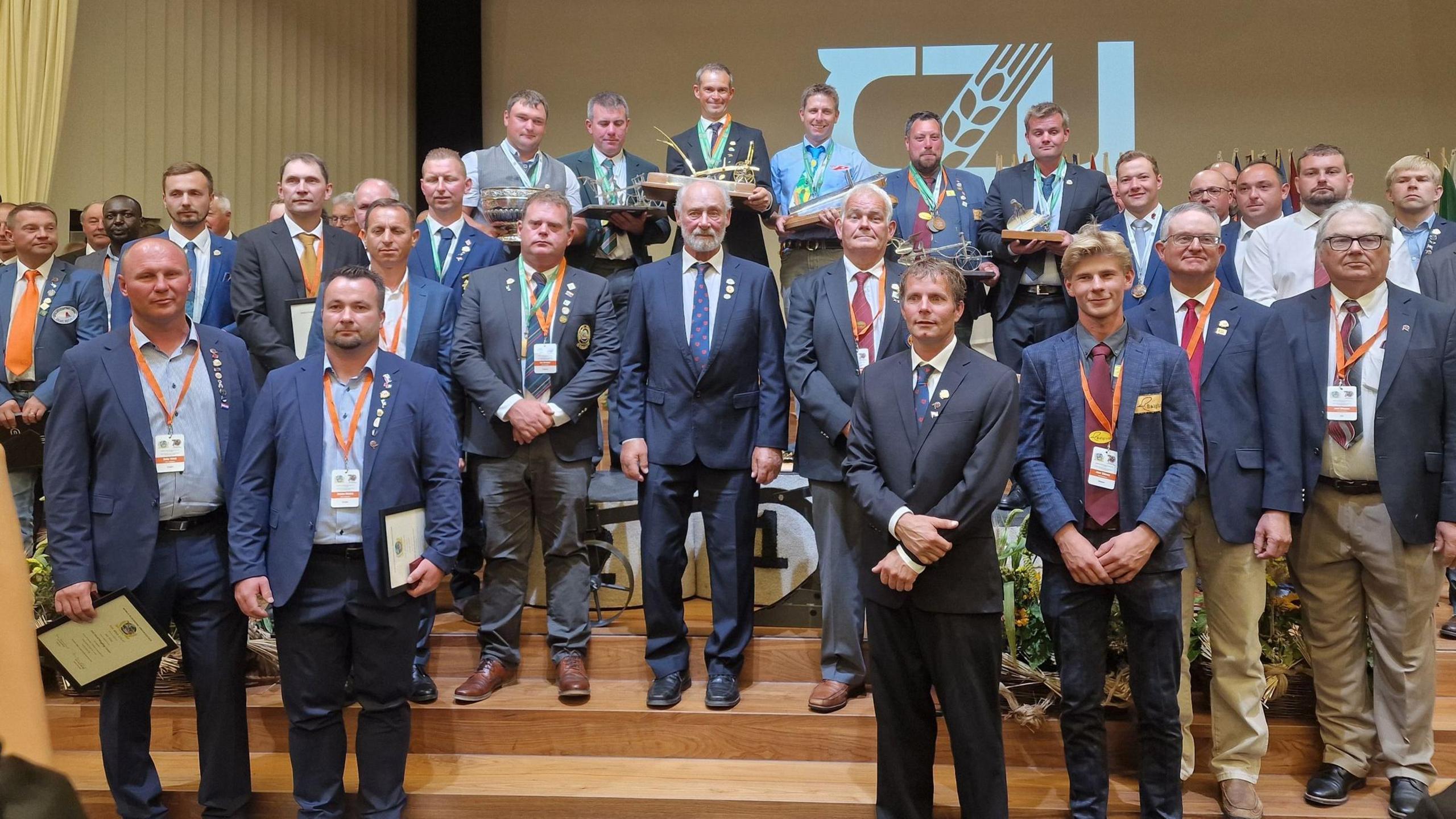 A mixed group of suited men stand on a stage, some holding trophies and wearing medals.