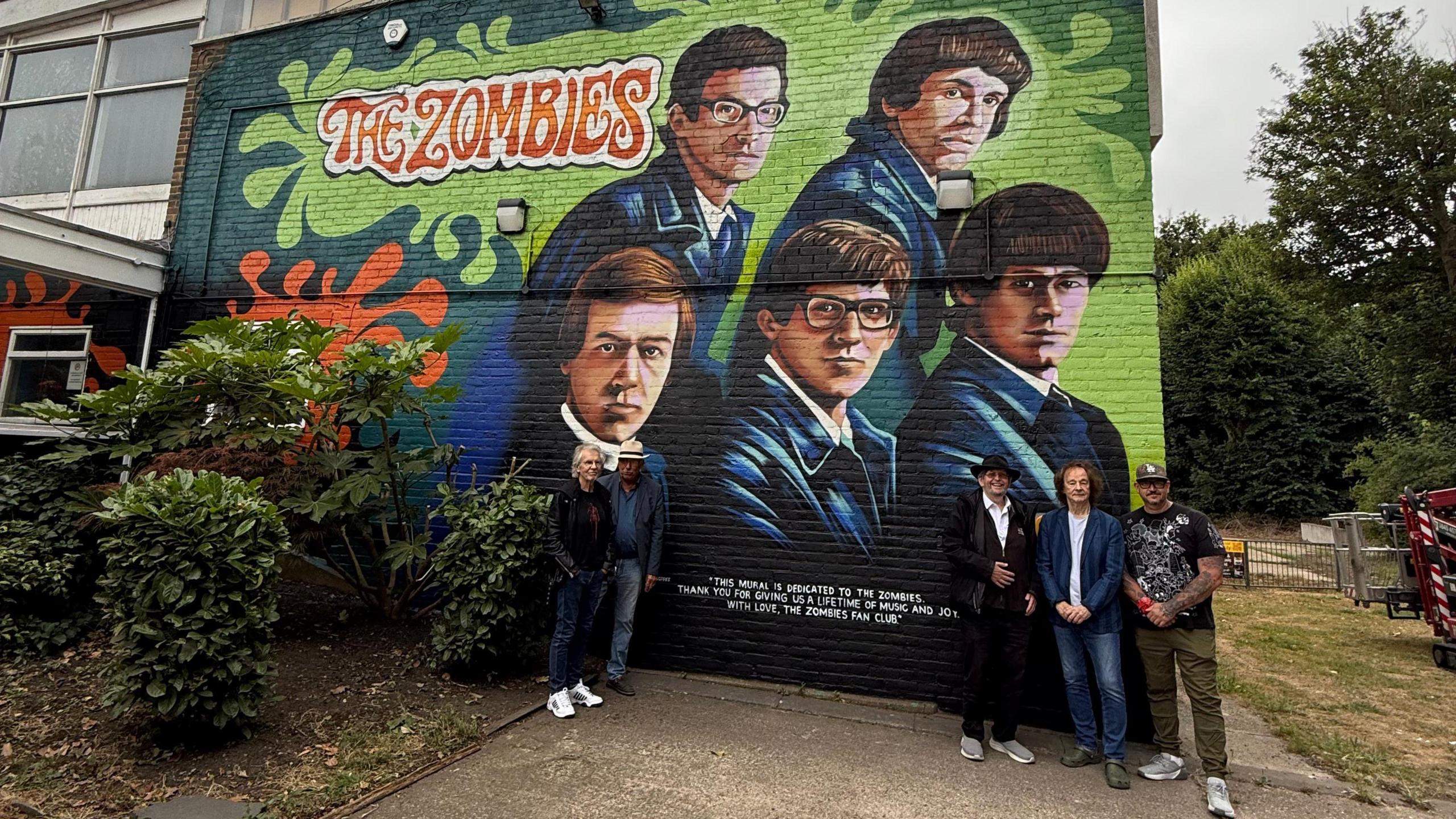 A mural depicting members of the music group The Zombies on the side of a building with members of the band standing in front of it. The mural is light green and dark green and depicts give men wearing blue velour or velvet jackets. They have 60s style hair and the text is written in a 1960s-style font.