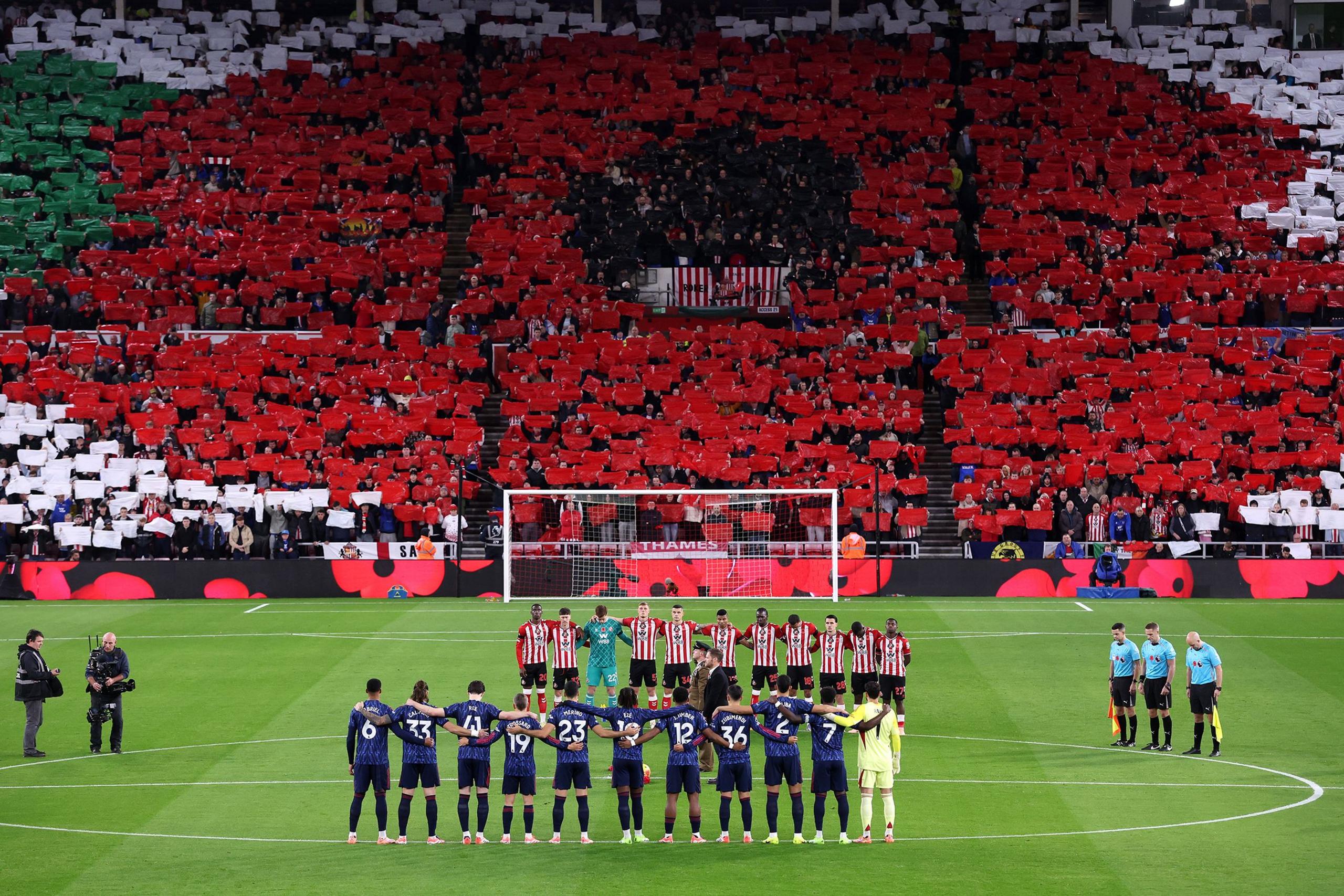 Two football teams and referees standing in formation on a green pitch for a pre-match tribute, with a stadium crowd holding red, white, and green cards arranged to form a giant poppy design under bright floodlights.