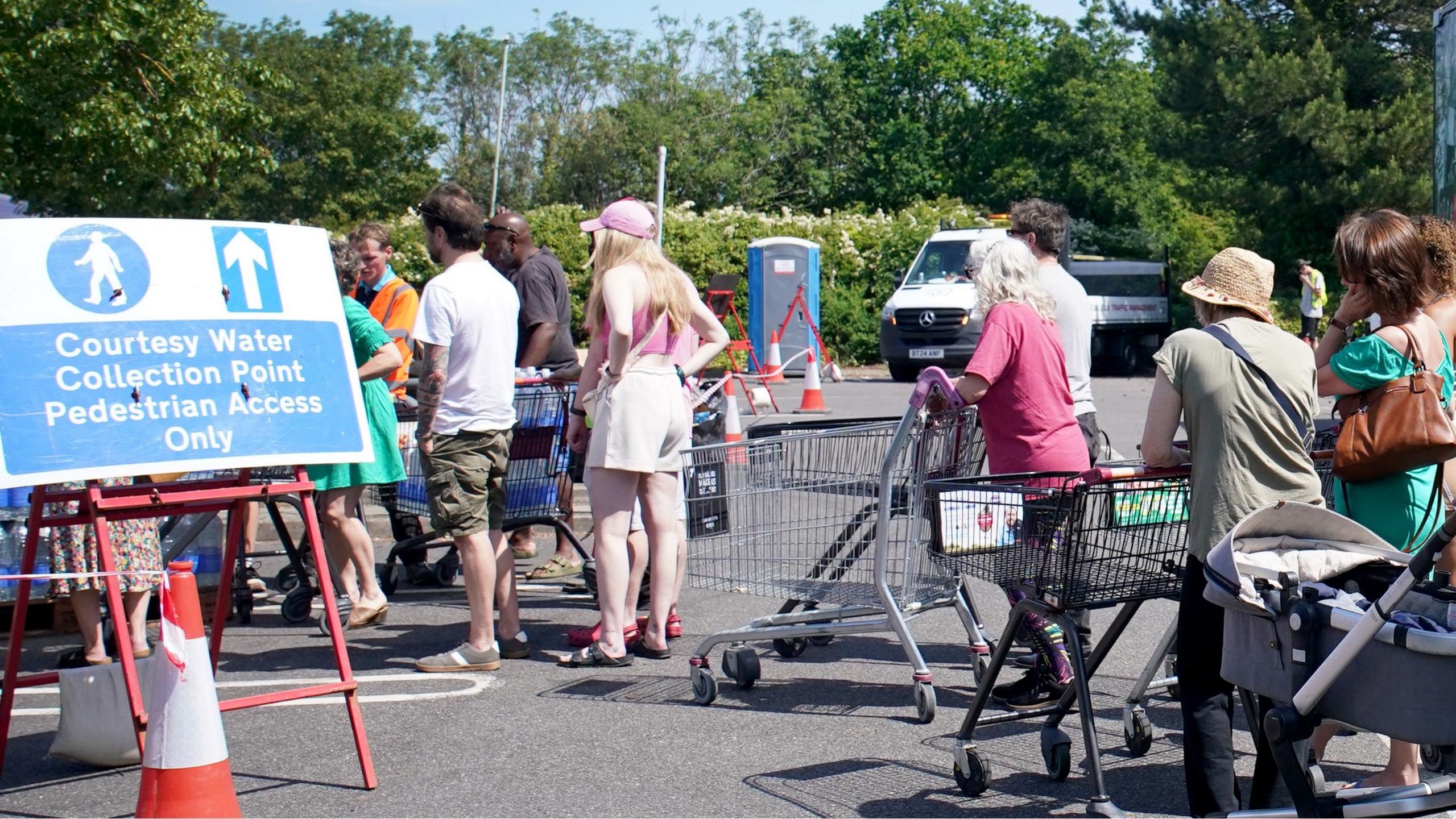 A line of people with trolleys queuing in the sunshine by a water collection point