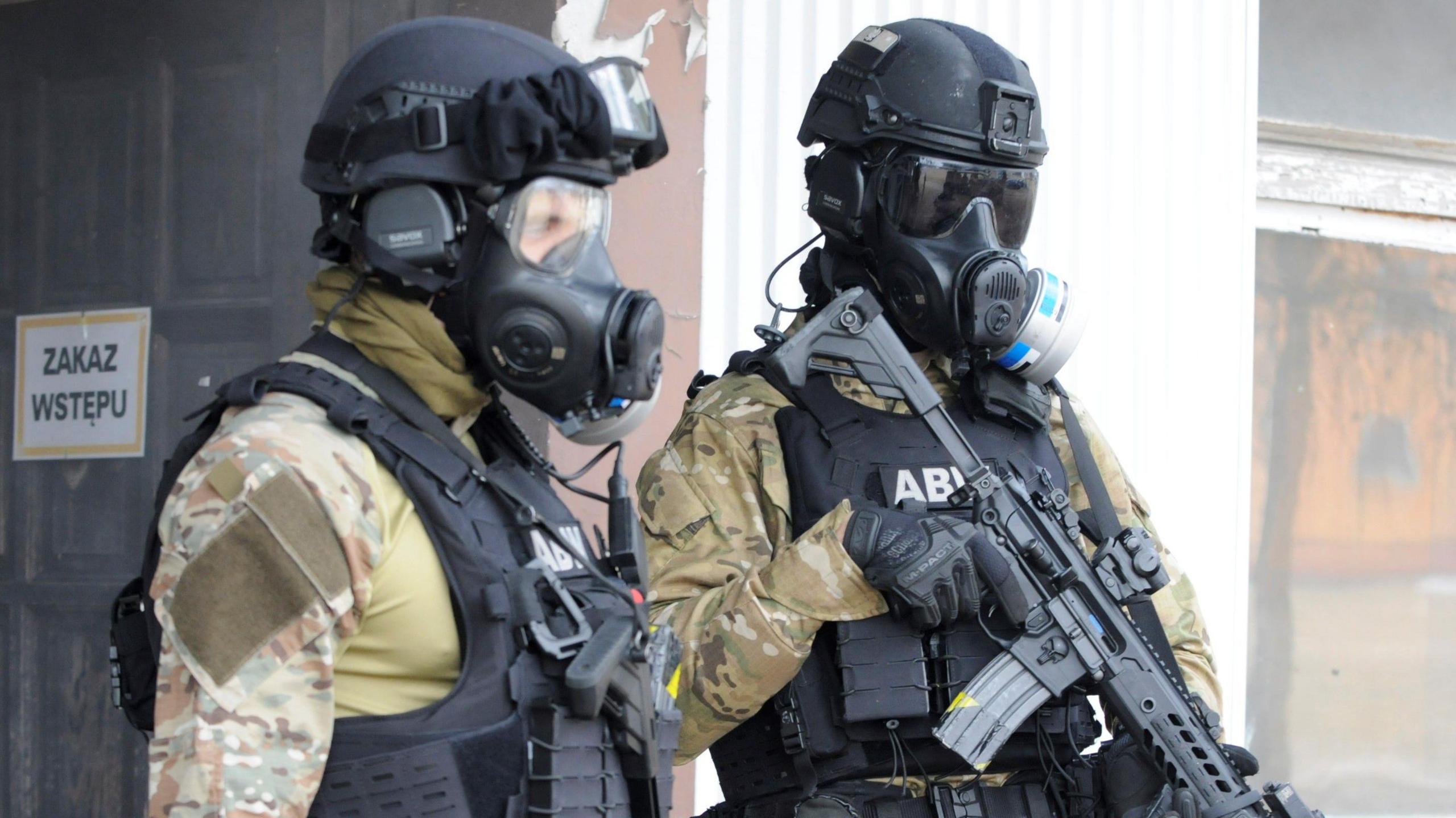 Two men wear gas masks and military uniform and carry guns