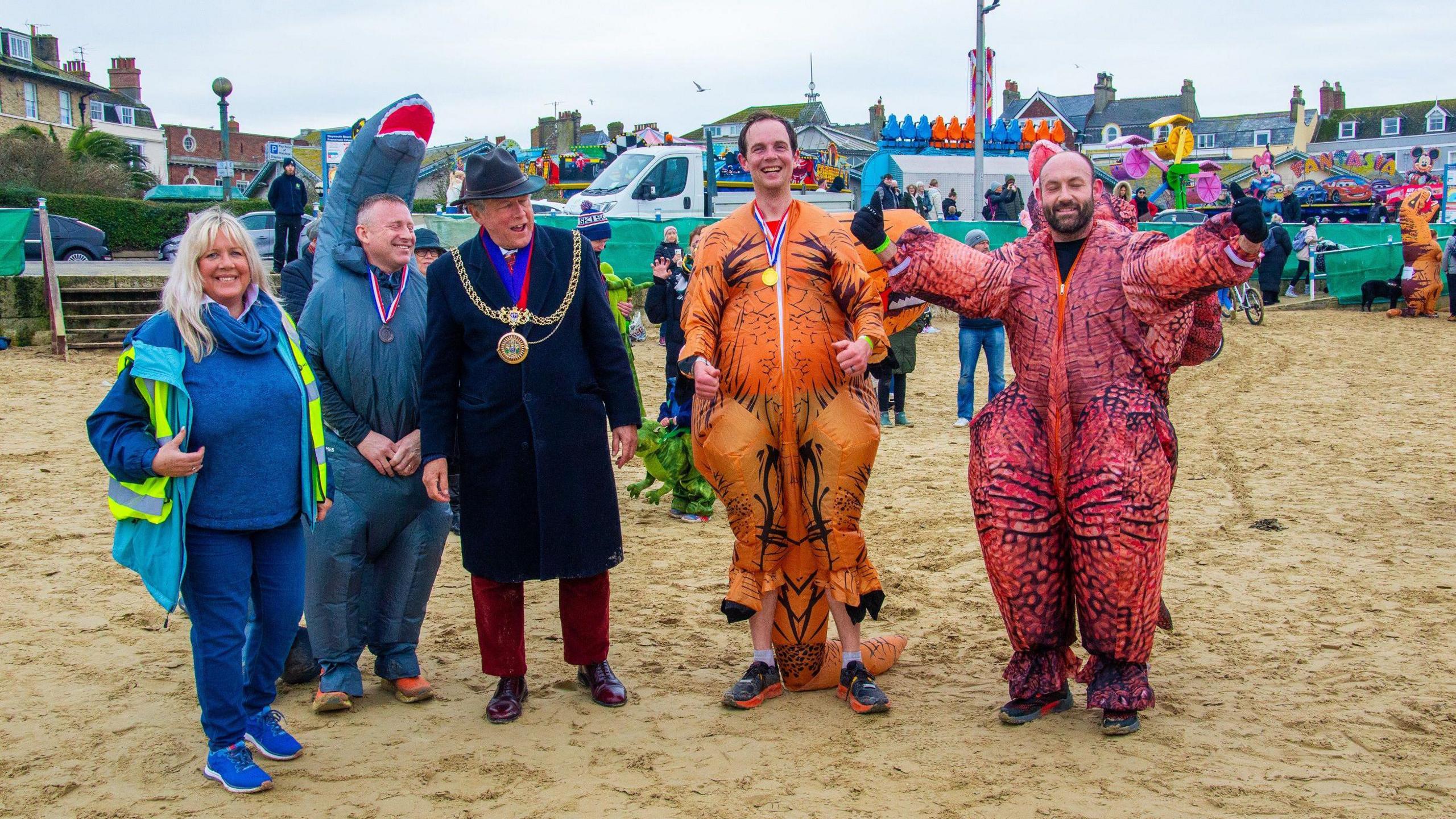 A man wearing ceremonial chains, a woman in a high-visibility jacket and three men wearing dinosaur costumes standing on a beach smiling at the camera.