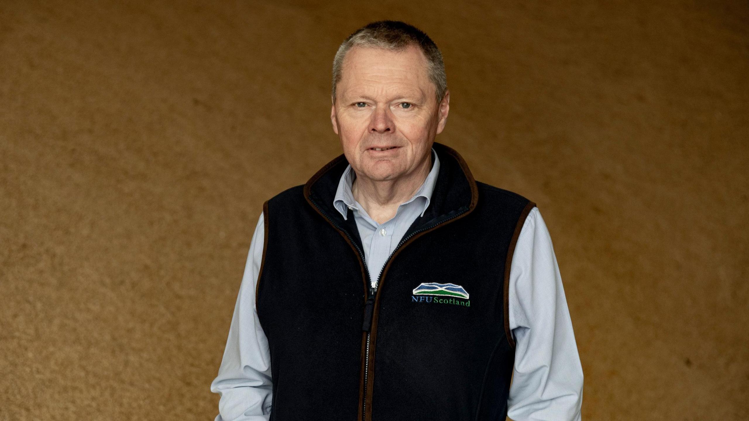 NFUS President, Andrew Connon standing infront of a pile of grain, wearing a blue gilet