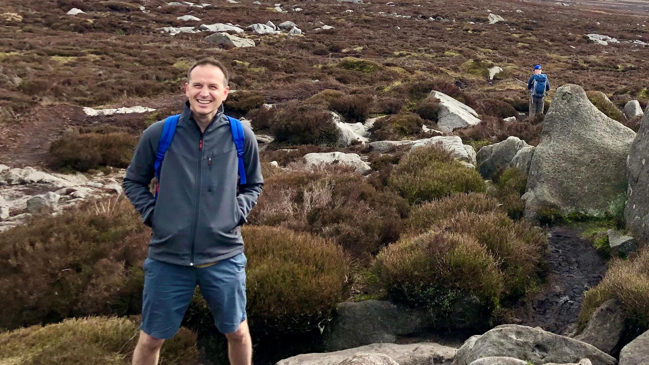 Tim Connors is standing on moorland with rocks around him. He is smiling at the camera and is wearing a dark jacket with the blue straps of a rucksack showing across his arms and dark blue shorts