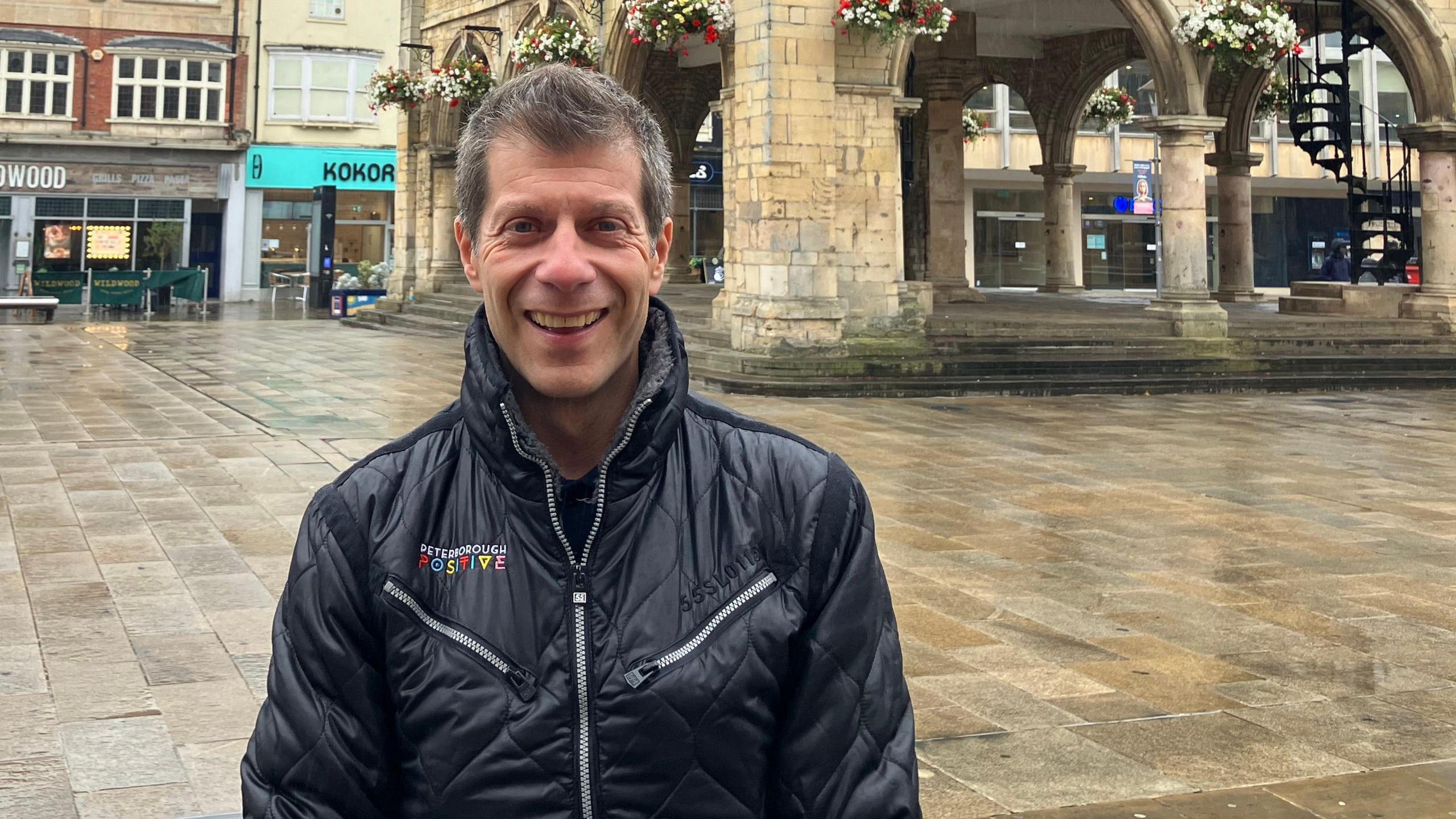 Pep wearing a black jacket smiling for the camera standing in front of a historic guildhall