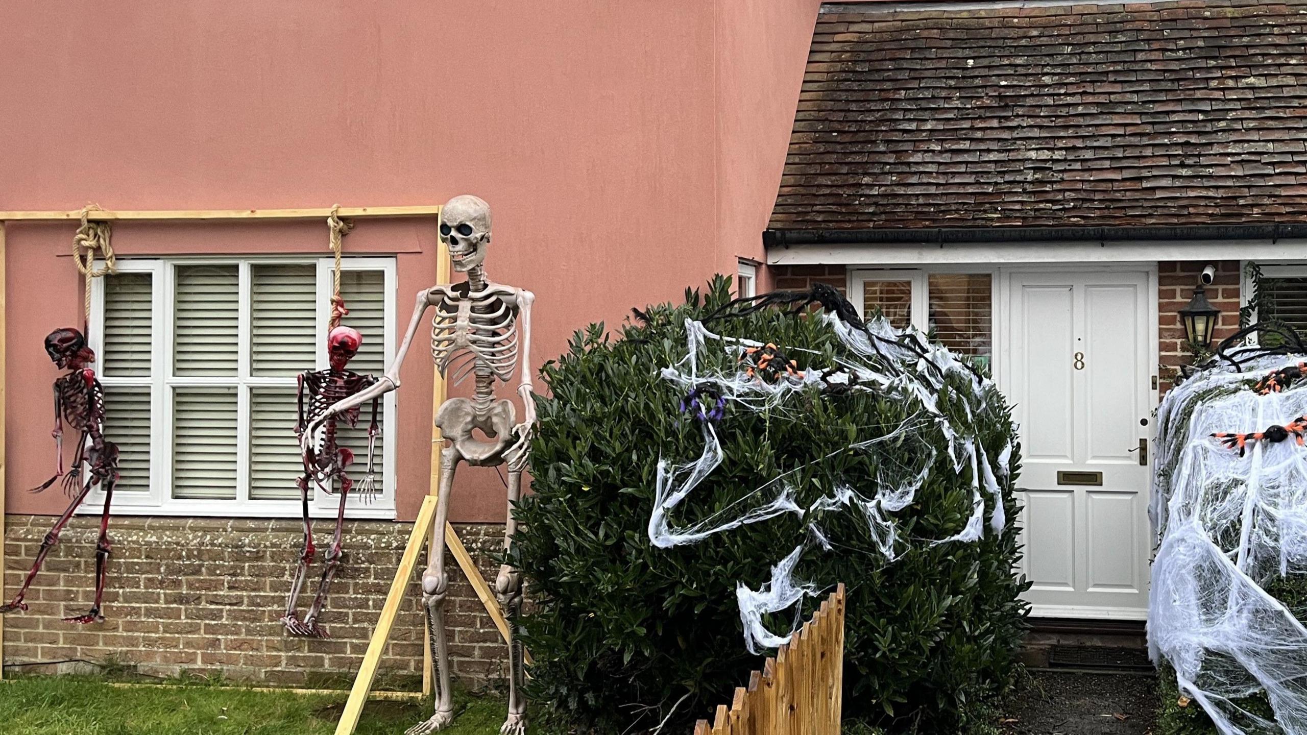 The front of a house. Bushes outside the front door have been covered in fake cobwebs and large spiders. A large skeleton stands to the left of the image next to one of the bushes. Two other smaller red skeletons hang from a wooden frame outside one of the house's front windows.