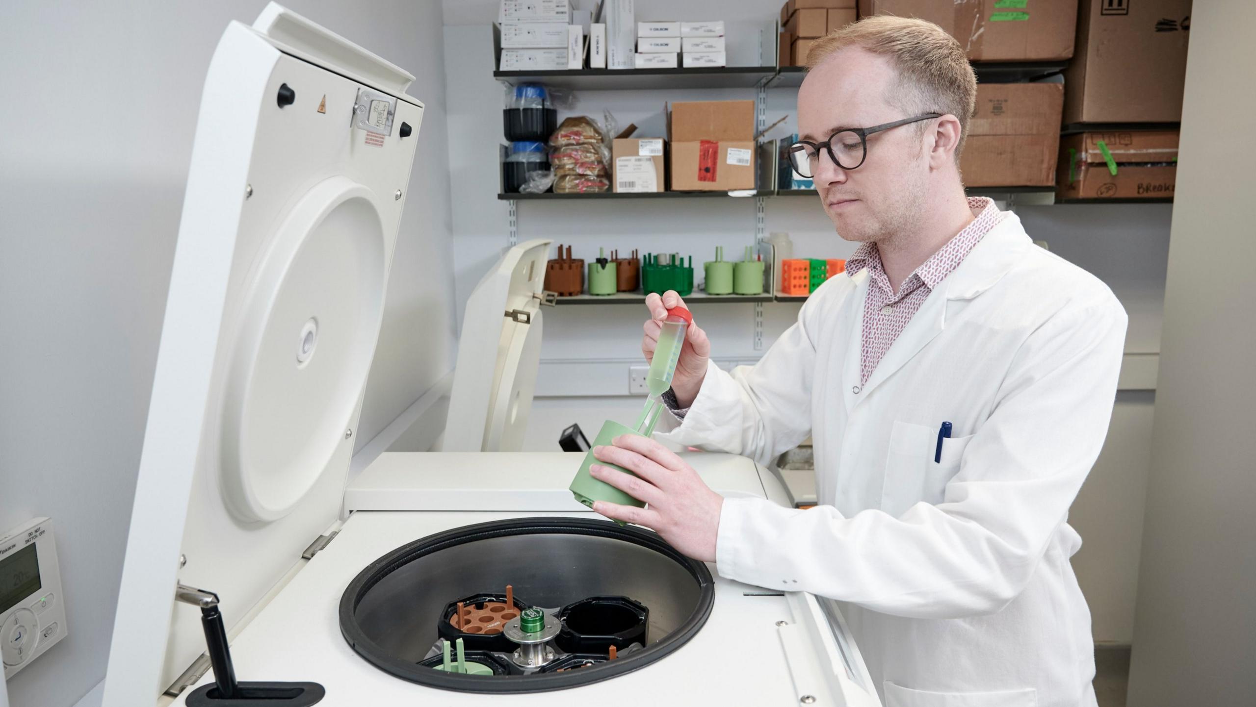 A scientist in a white coat standing over a machine, adding a solution into a container from a vial