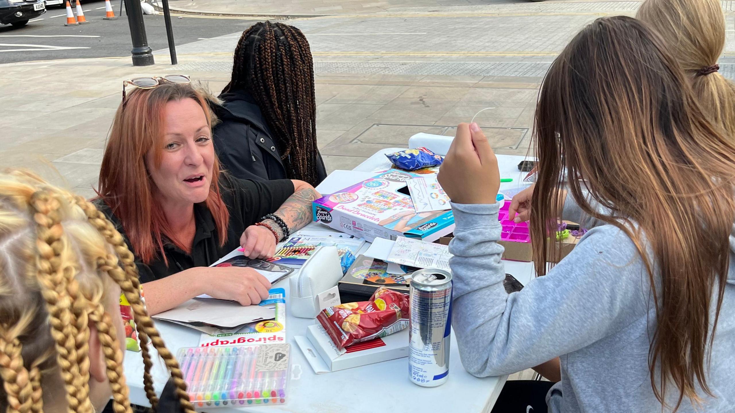 Katy Shroff and three anonymous teenage girls at a table in Broad Street, March. The table is covered in colouring pens and pencils, plus a couple of packets of crisps. Katy is on the far left, has crouched down at the table and looking towards the viewer and smiling. She has reddish shoulder-length hair, with her glasses perched on her head and is wearing a black top. The three girls all have long hair and have turned their backs to the viewer.