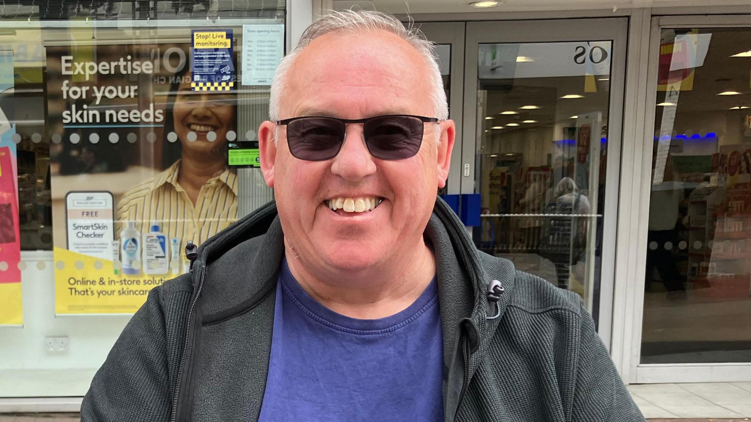 Keith Horner has white hair, darkly tinted glasses, a blue t-shirt and a darkly-coloured hooded top. He is sitting or standing in the high street, and a shop frontage is behind him.