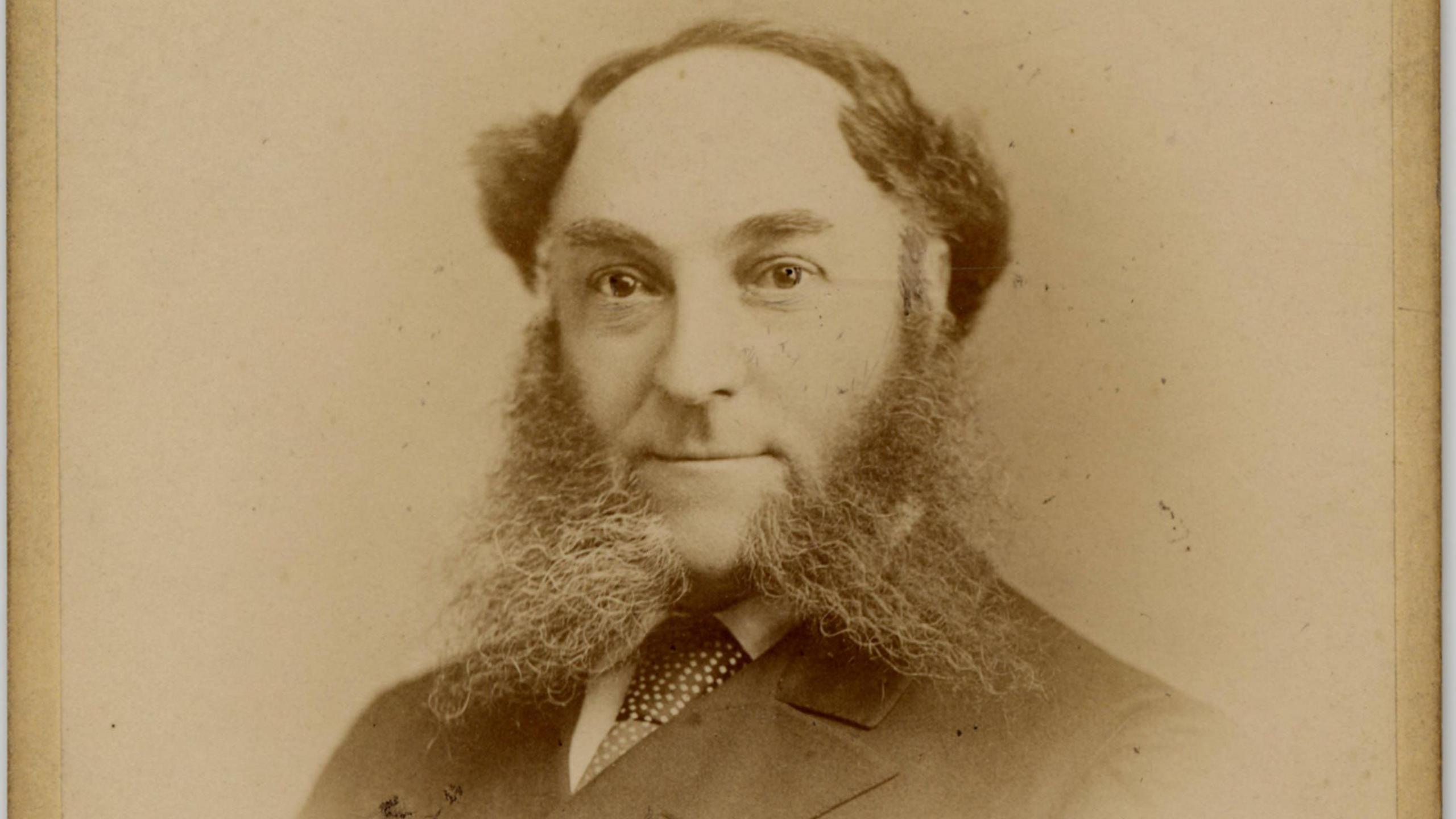 Sepia photo of William Whiteley, 1907. A balding man wearing a suit in a formal portrait. He has massive whiskers on each side of his chin .