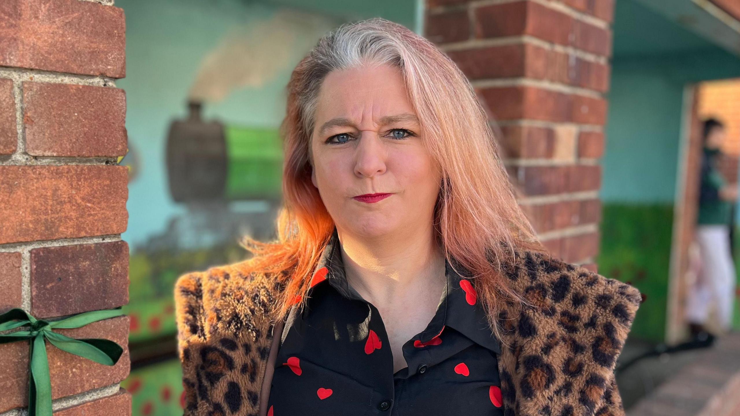 Katie Hart, a woman who is wearing a black blouse with red love hearts on it with a leopard print fur coat over the top. She is looking directly at the camera and is standing at the Sheringham bus shelter which has a painted mural in it.