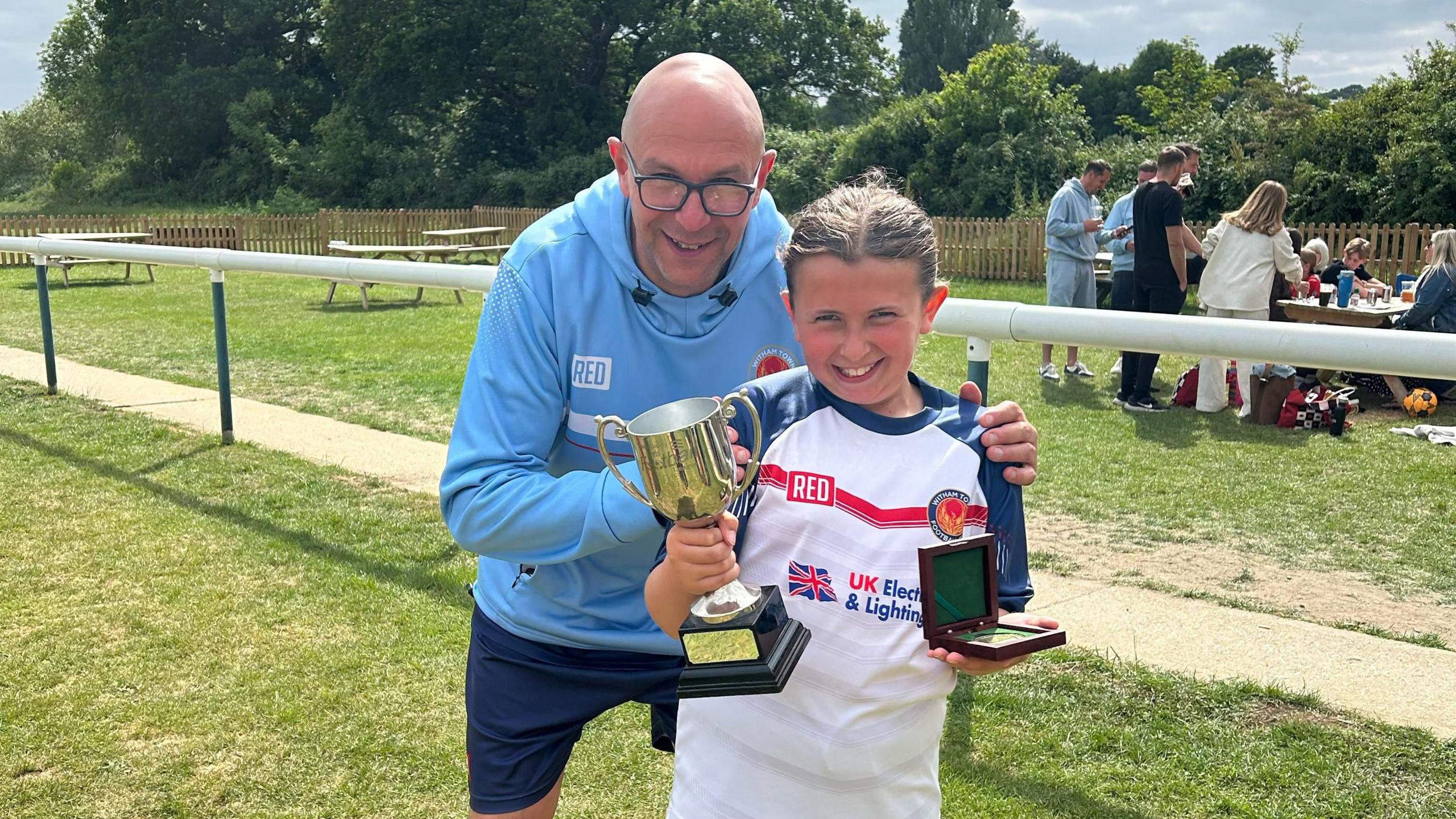 A girl holding a trophy and a medal box with a football top on and a man standing behind her looking proud