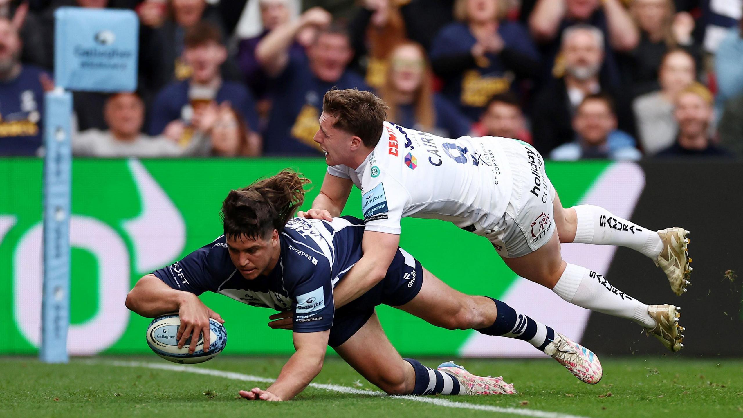 Benhard Janse van Rensburg of Bristol Bears dives over the line to score a try as Tom Cairns of Exeter Chiefs tries to tackle him. In the background fans can be seen celebrating