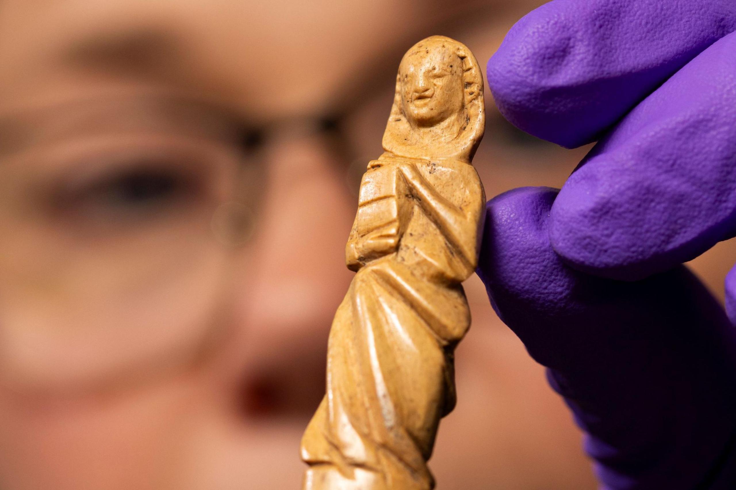 A close up of the gravoir, an ornately carved piece of red deer antler. The carving is of what looks like a hooded figure holding a book. Dr Alice Blackwell is holding the gravoir in protective purple gloves. Dr Blackwell's face is just out of focus.