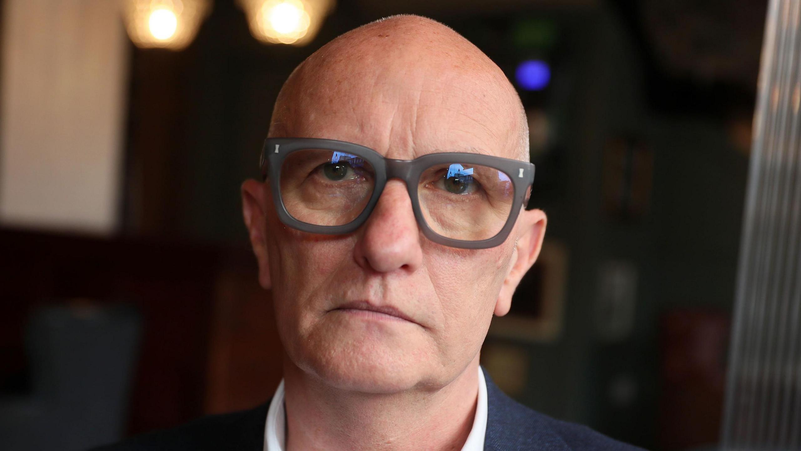 Colin Neill - A bald man wears a pair of large, black square-framed glasses, a navy suit blazer and a white collared shirt. He is looking at the camera and the background behind him is blurred.