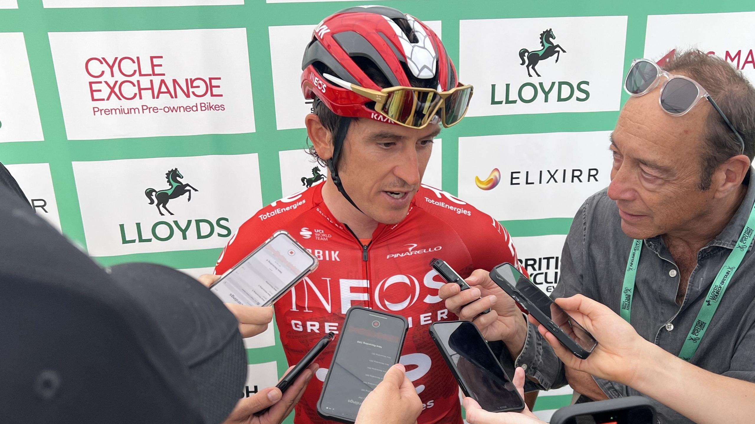 Cyclist Geraint Thomas speaks to reporters who hold phones and microphones close to his face. He wears a red cycling helmet with gold sunglasses that rest on top. He is also wearing a red cycling jersey. 