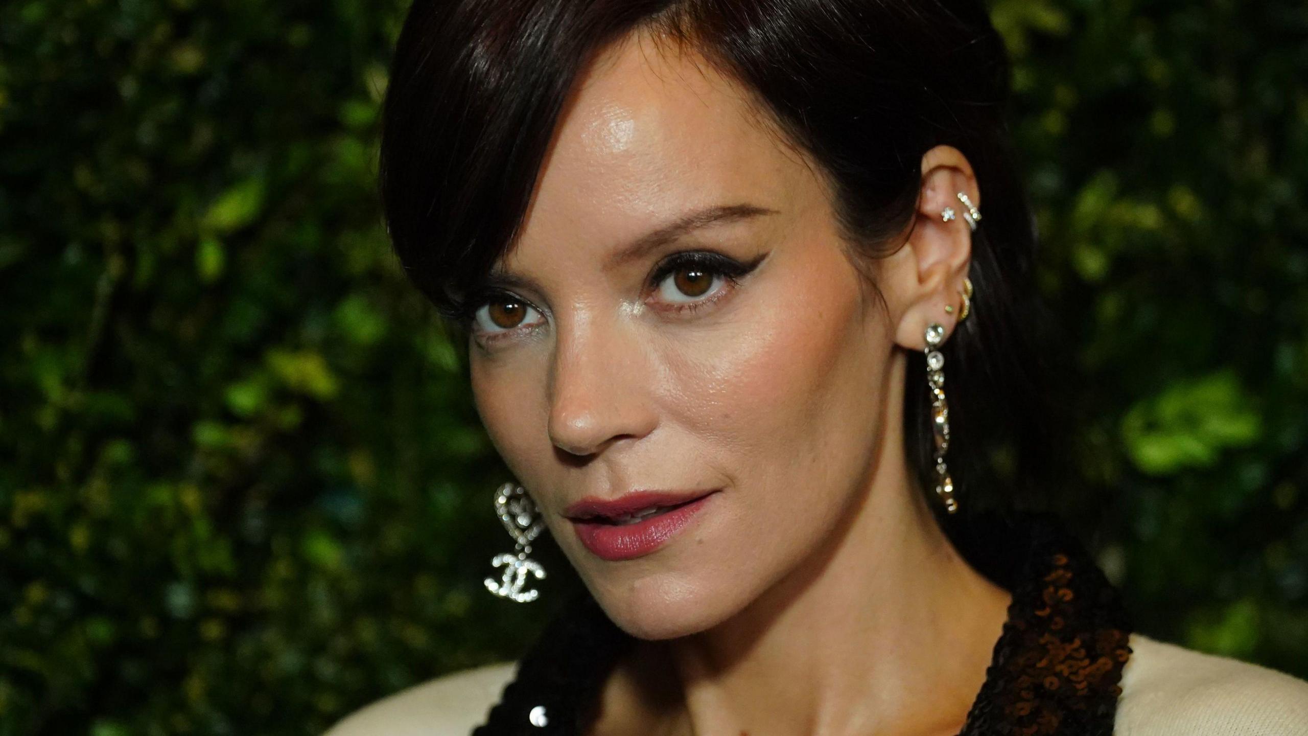 Lily Allen, pictured with black hair in an up do with a side fringe, winged eye liner and several diamond earrings