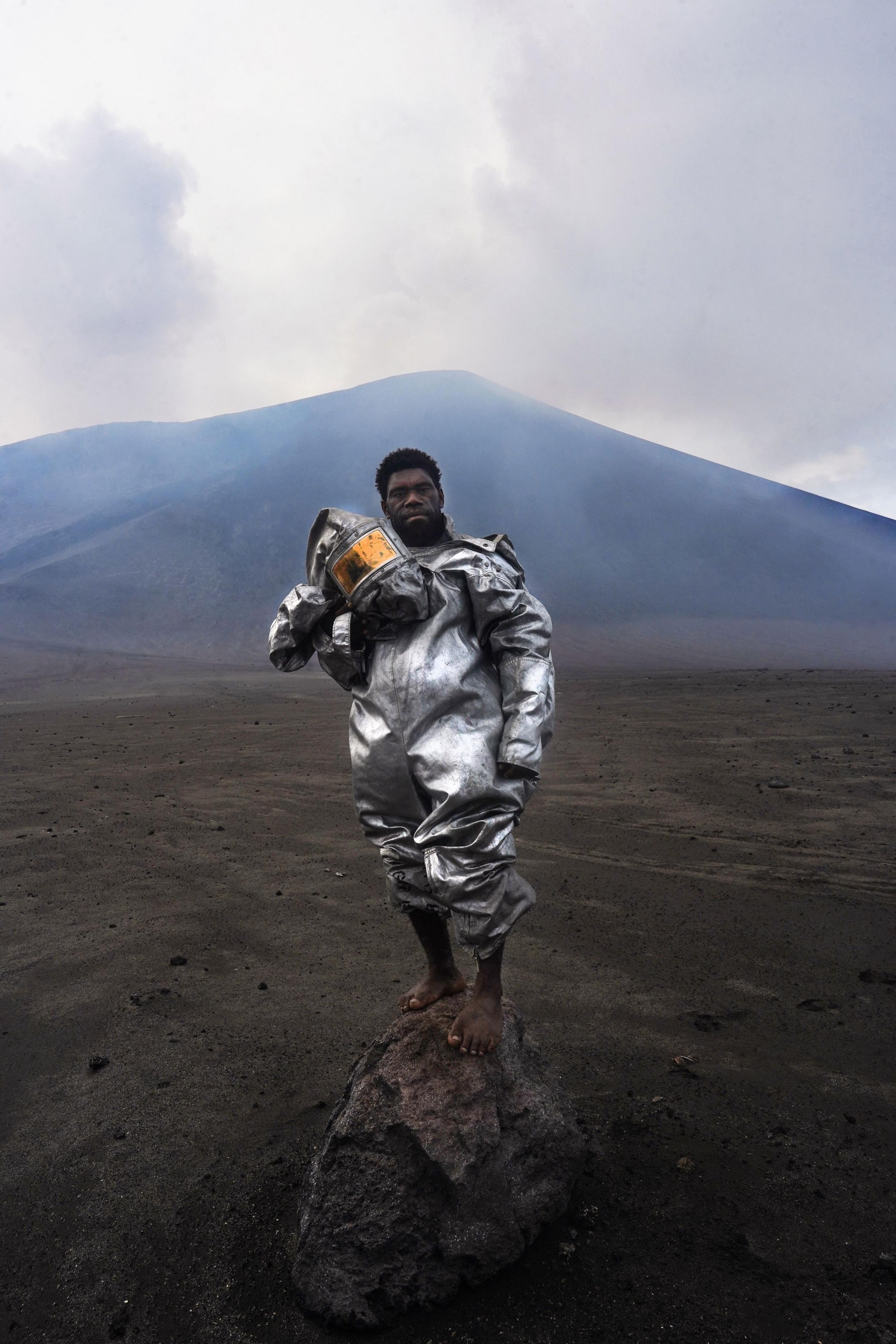 a man standing on a rock in a silver suit and holding a silver helmet, with a volcano behind him
