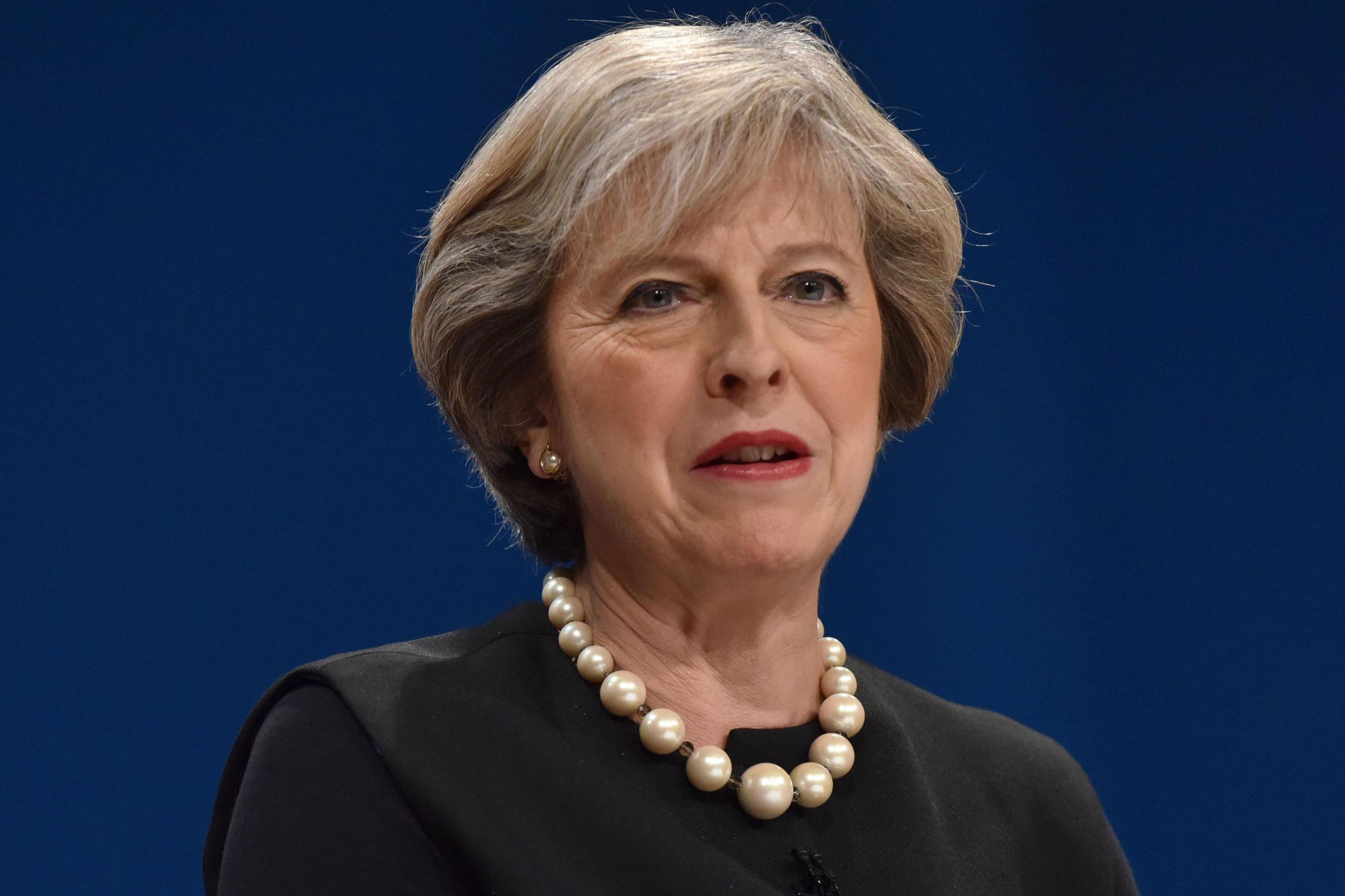 Former conservative Prime Minister against a blue background, wearing a pearl necklace and black jacket.