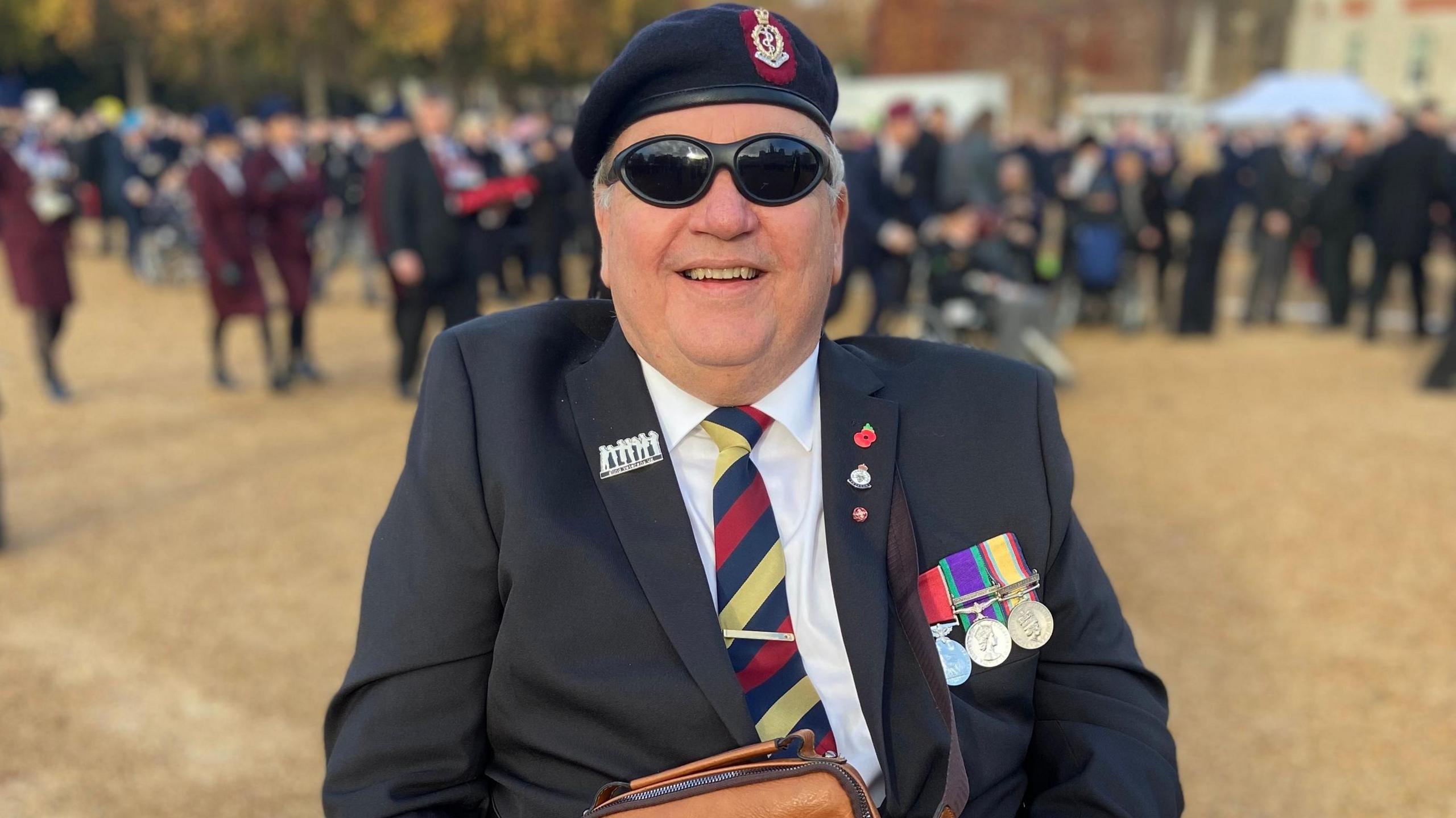 John Hardy. He is pictured outside at a Remembrance Sunday service. He is wearing a suit and his medals are pinned to the jacket. He is also wearing dark sunglasses as he is blind. He is smiling at the camera. A crowd of people can be seen in the distance behind him.