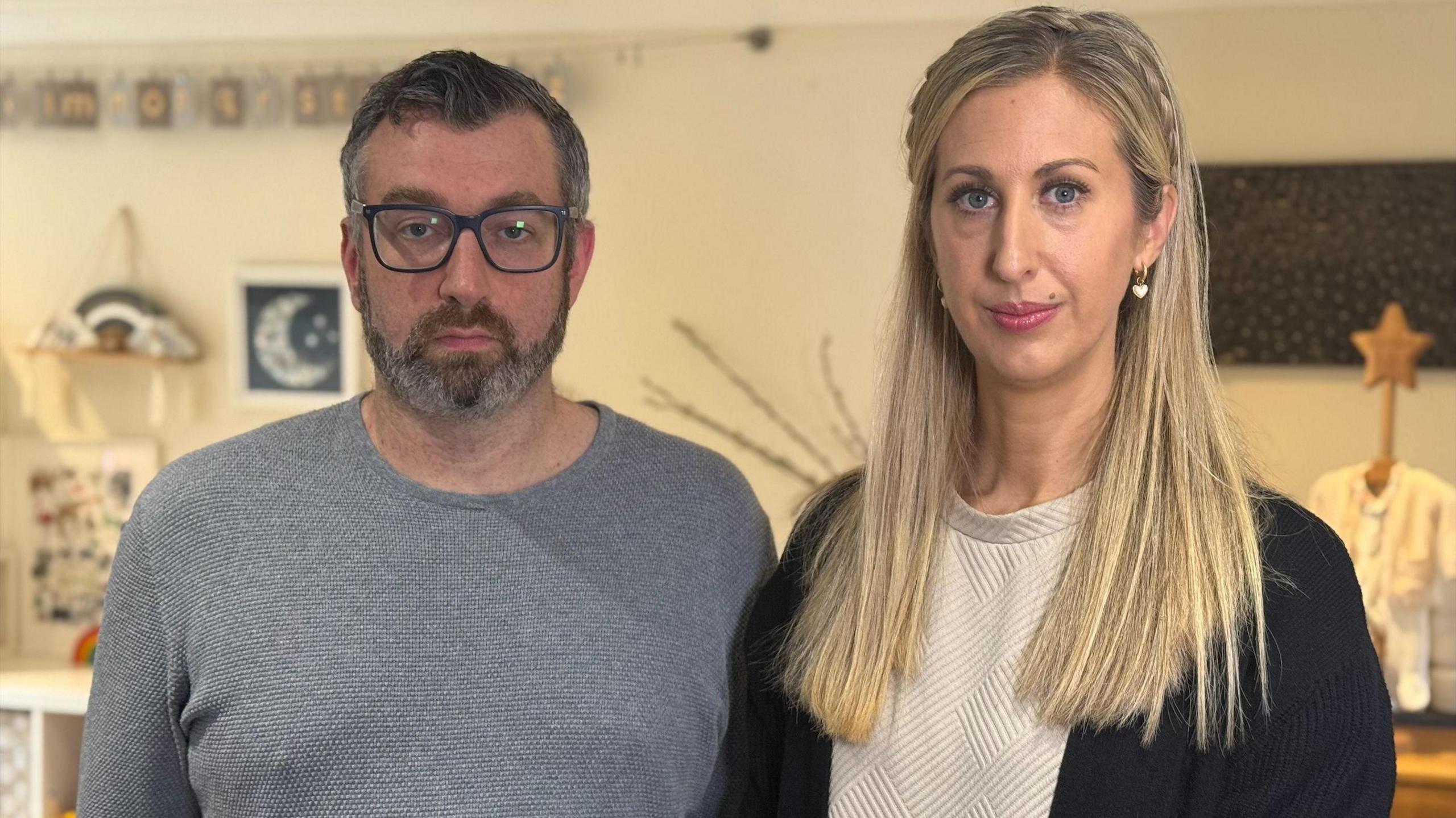 A man and a woman stand in a baby's nursery, facing the camera with serious expressions. Dan, who has a beard, is wearing a grey jumper and glasses. Fiona has a white top and a black cardigan and wears her straight blonde hair in a plait.