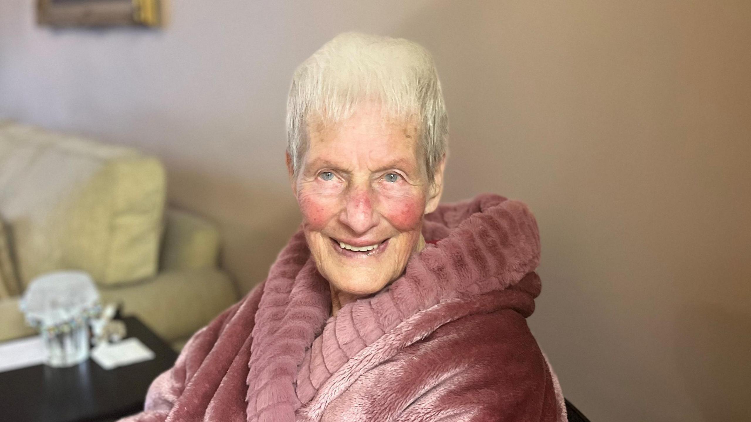 Betty McKenzie is sitting in a living room. She has short grey hair and red cheeks. She is wearing a pink dressing gown. She has a smile on her face.