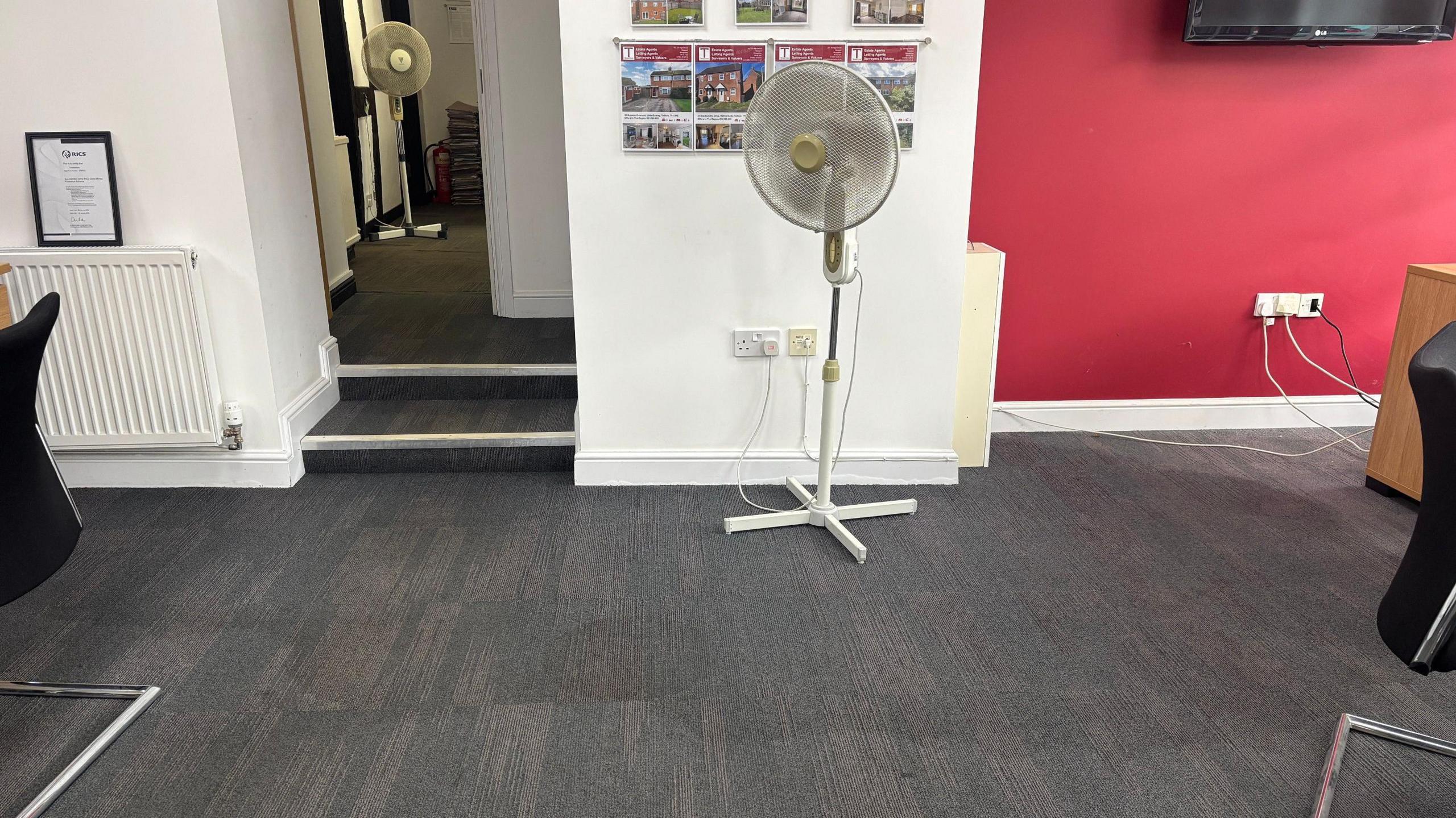 A white standing fan in a room with a large wet patch on the floor. In the background is another white fan