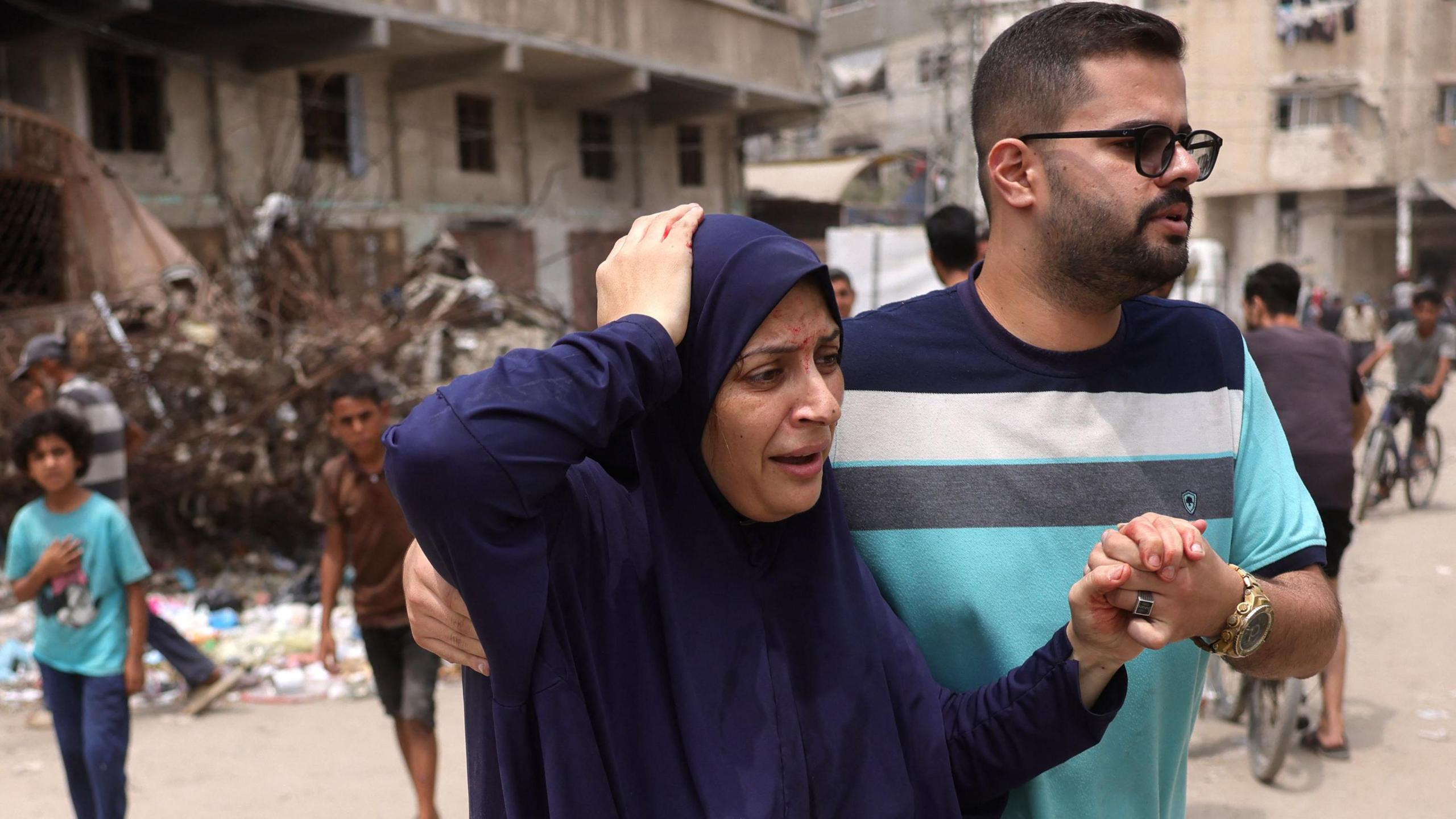 An injured Palestinian woman wearing a blue robe holds her hand to the top of her head looking in pain as a man helps her by holding her left hand with his and the right hand behind her back. He is wearing a striped light blue, grey, white and navy blue shirt, has a stubble beard and is wearing glasses. Behind them there are two children and a building whose ground floors appears to have been hit