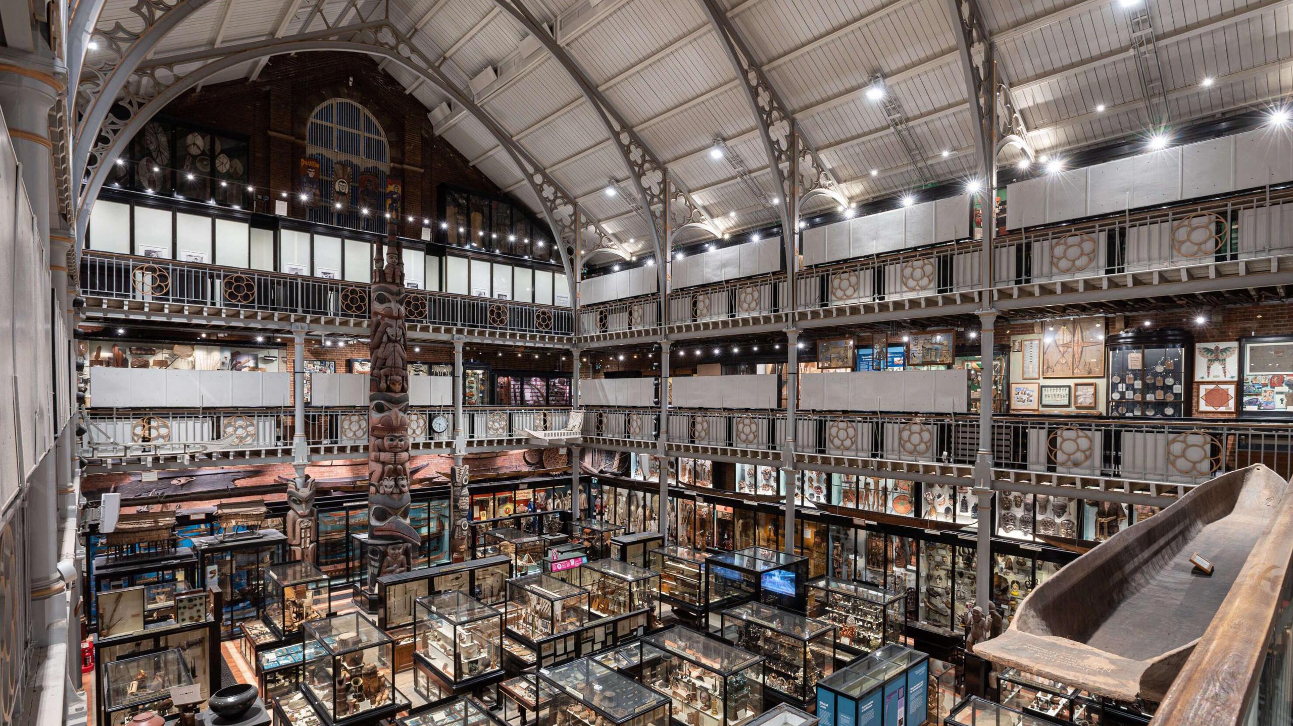 A view of the Pitt Rivers Museum. On the floor are lots of glass casses containing artefacts. A large totem pole soars above the ground. There are various floors and a white chapel-like roof. 