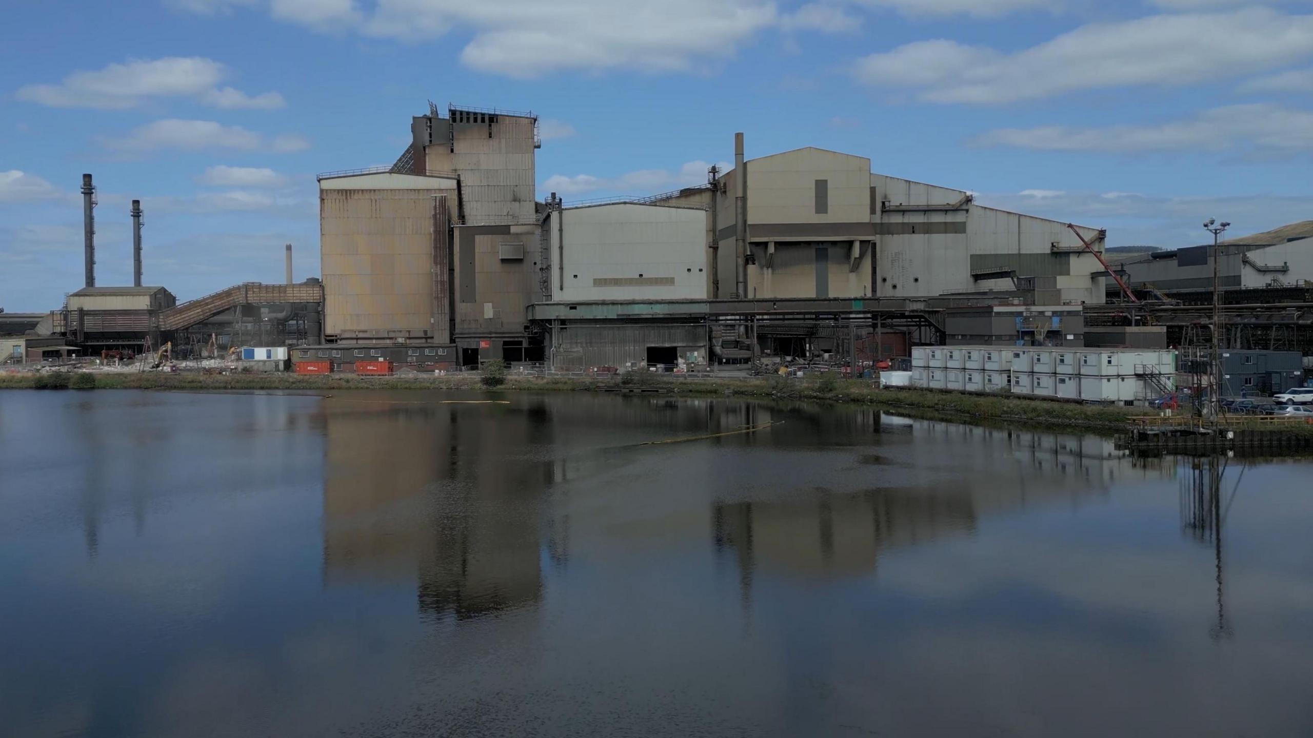 An industrial steelmaking site in Port Talbot, South Wales, seen from across a calm body of water. Several large buildings dominate the scene, including tall chimneys and metal-clad structures. Construction materials and equipment are visible near the buildings, suggesting ongoing work. The buildings are reflected clearly in the water, and the sky above is partly cloudy with patches of blue.