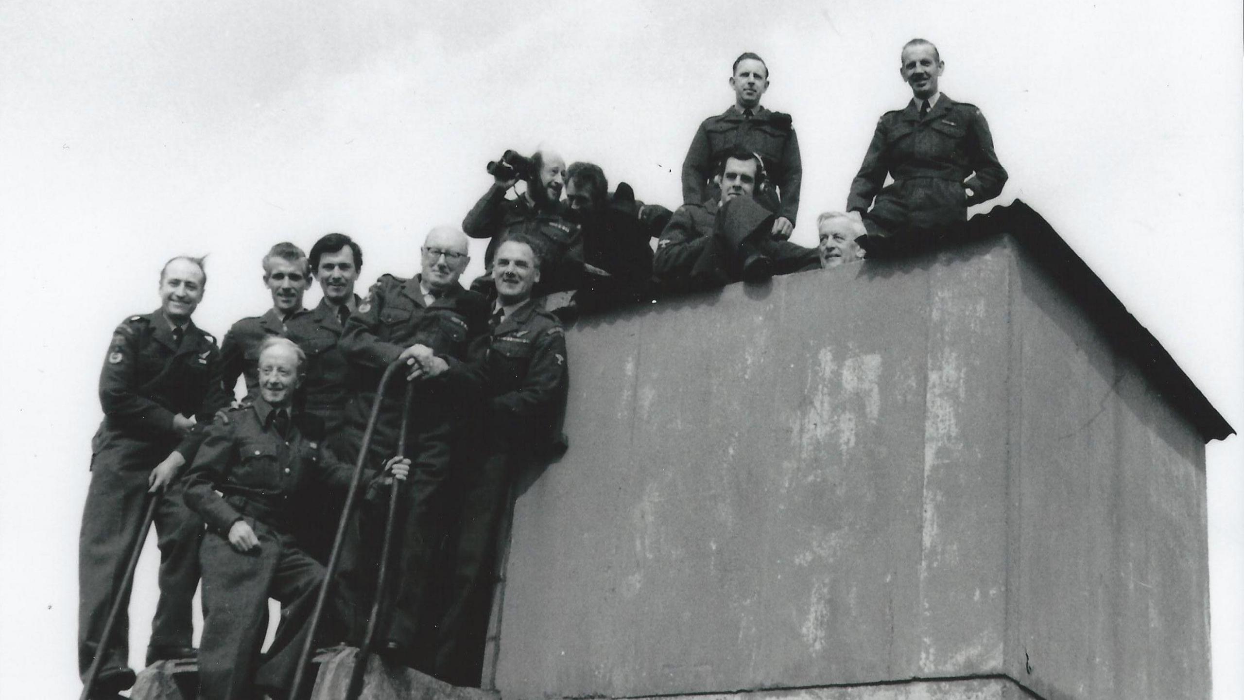 A black and white image of group of men in military uniform. They are gathered on the top steps of a ladder and the roof of a concrete structure. The man in the centre of the group holds a pair of binoculars.