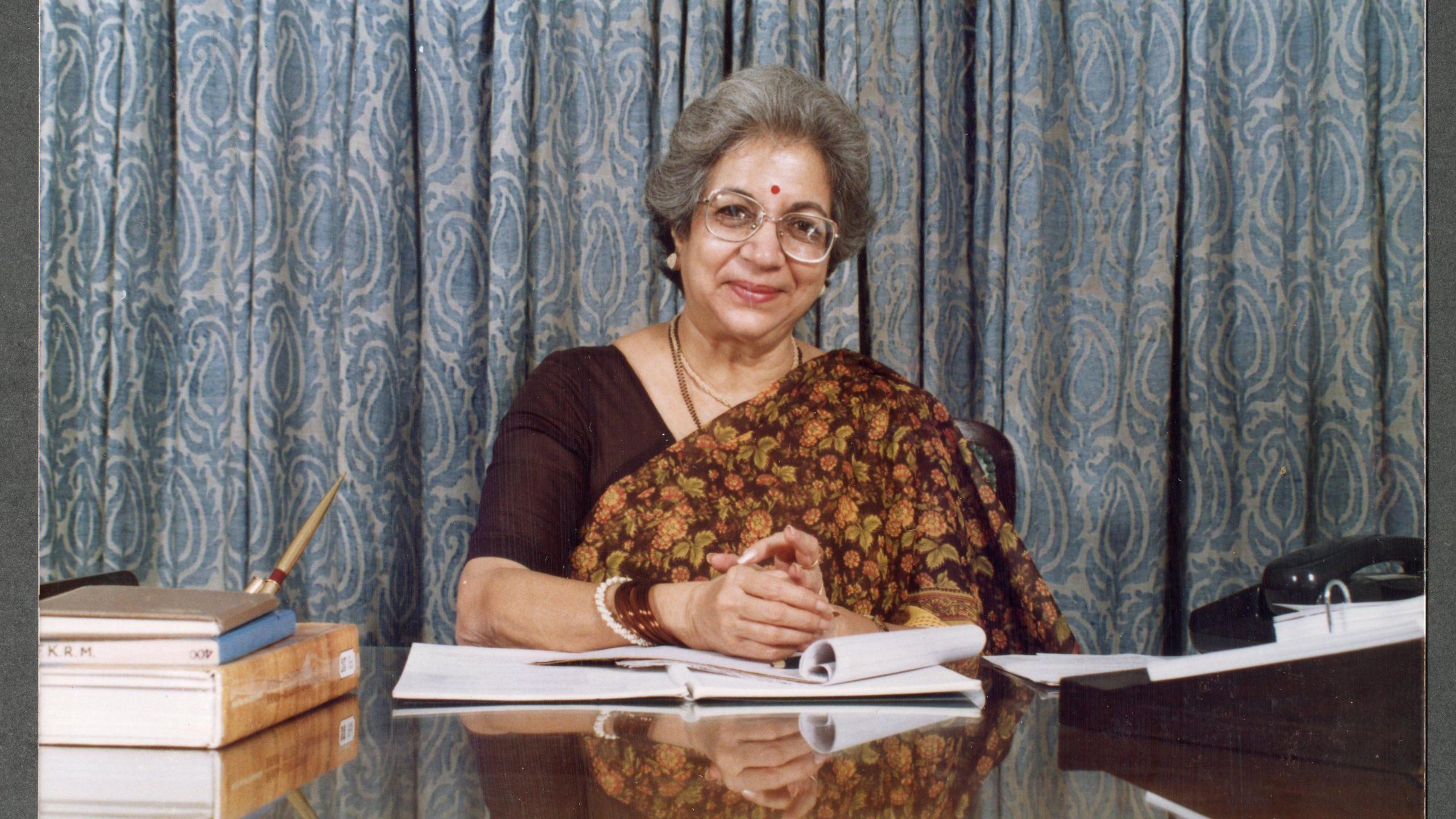 A photo of Vijaya Mehta sitting at her desk in her office and writing in a book.