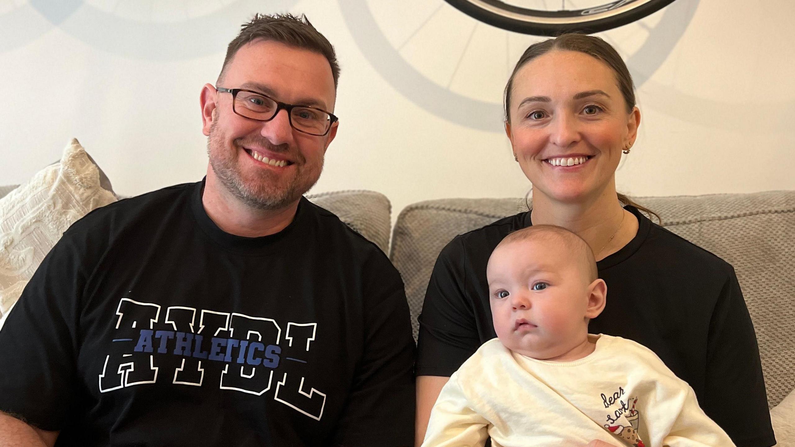 Chris and Jade are sitting on a couch holding baby Arwell who is dressed in a cream-colored top and red plaid pants. Behind them, two bicycles are mounted on the wall. Cushions and a throw blanket are visible on the couch.
