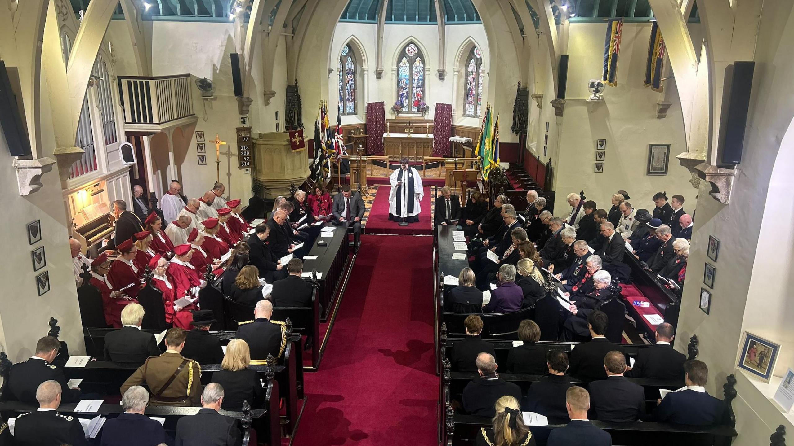 The interior of a church, which has people in dark clothing sitting in wooden pews with their heads bowed. There is a red carpet along the centre isle and stained glass windows at the far end.