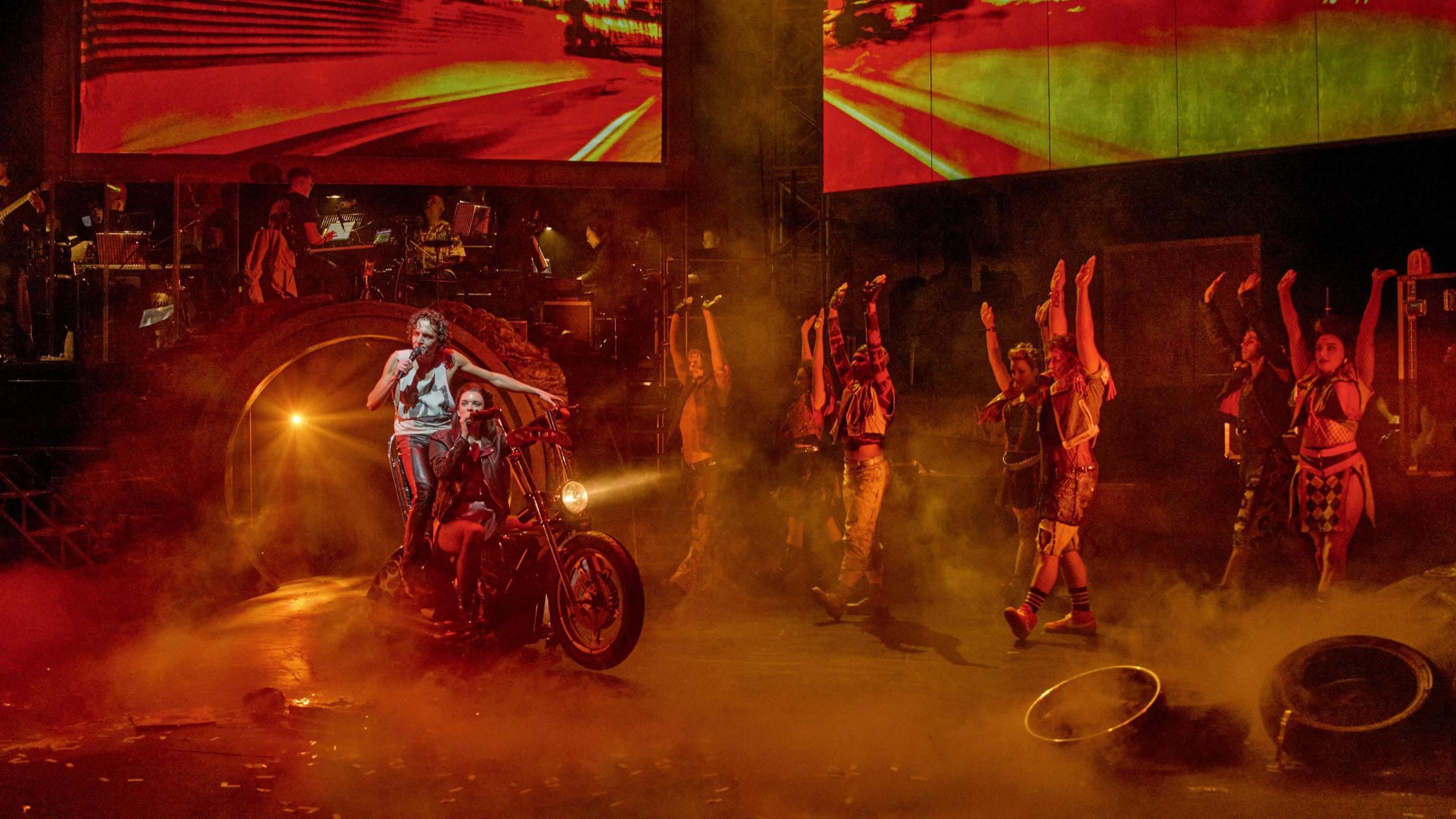 A wide image of cast on stage during Bat out of Hell at Bristol Hippodrome. Two actors are riding a Harley Davidson while other cast members, dressed in punk-style outfits, are dancing in line. The stage is lit up with a reddish light and there are clouds of smoke going across it