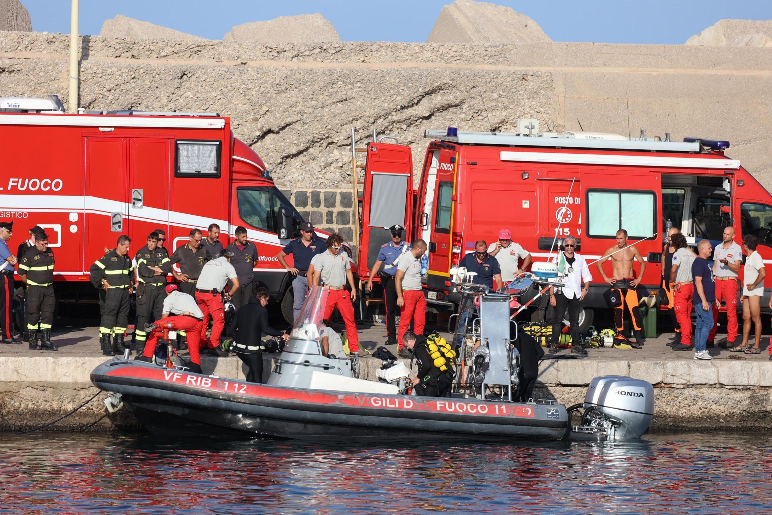 Two red emergency vehicles are visible at the dockside in Italy. A large number of rescue crew are also at the scene, with a life boat sitting just off the dock