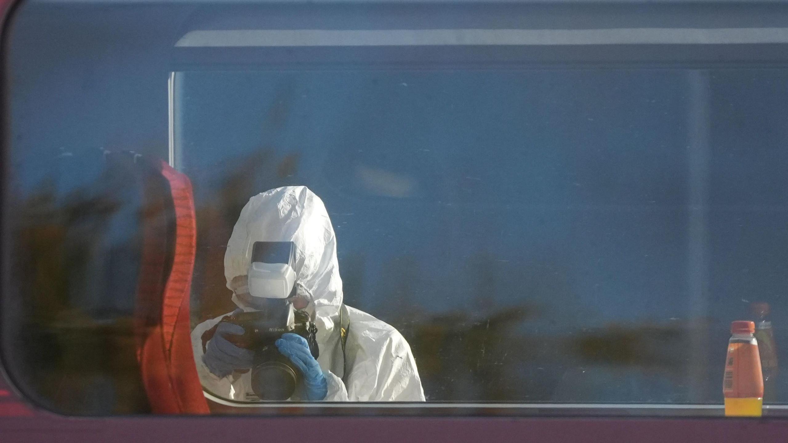 A person is stood in a train taking a picture of one of the red seats. They are wearing a white hazardous suit. 