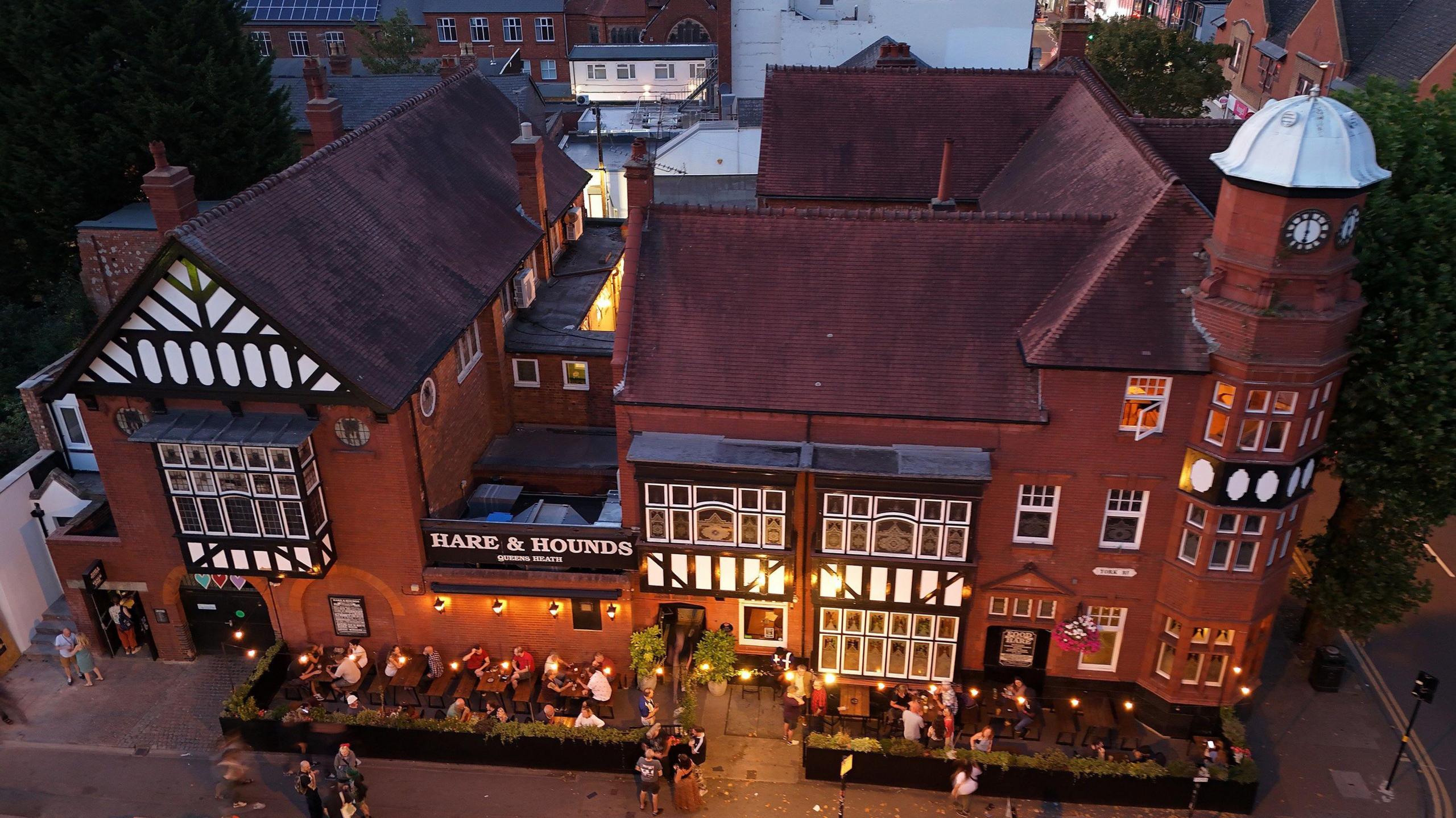 Aerial shot of the Hare & Hounds pub. It is a red brick building with tudor-style timber beams. Customers can be seen sitting at outdoor seating areas.