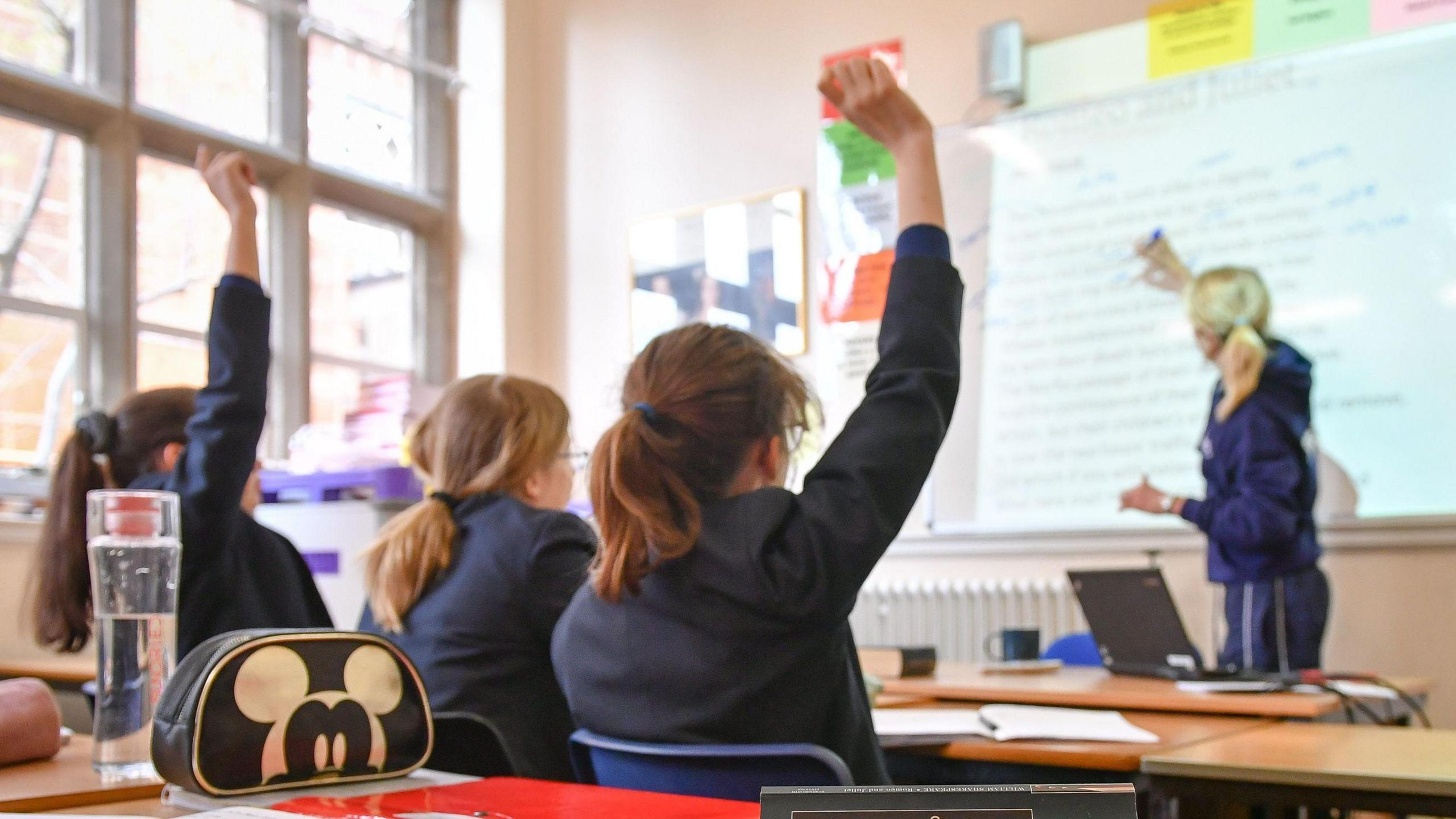 The backs of three schoolgirls in a classroom with two of them raising their hands and looking at a blonde teacher wearing a hoodie who is looking at an interactive large teaching screen.