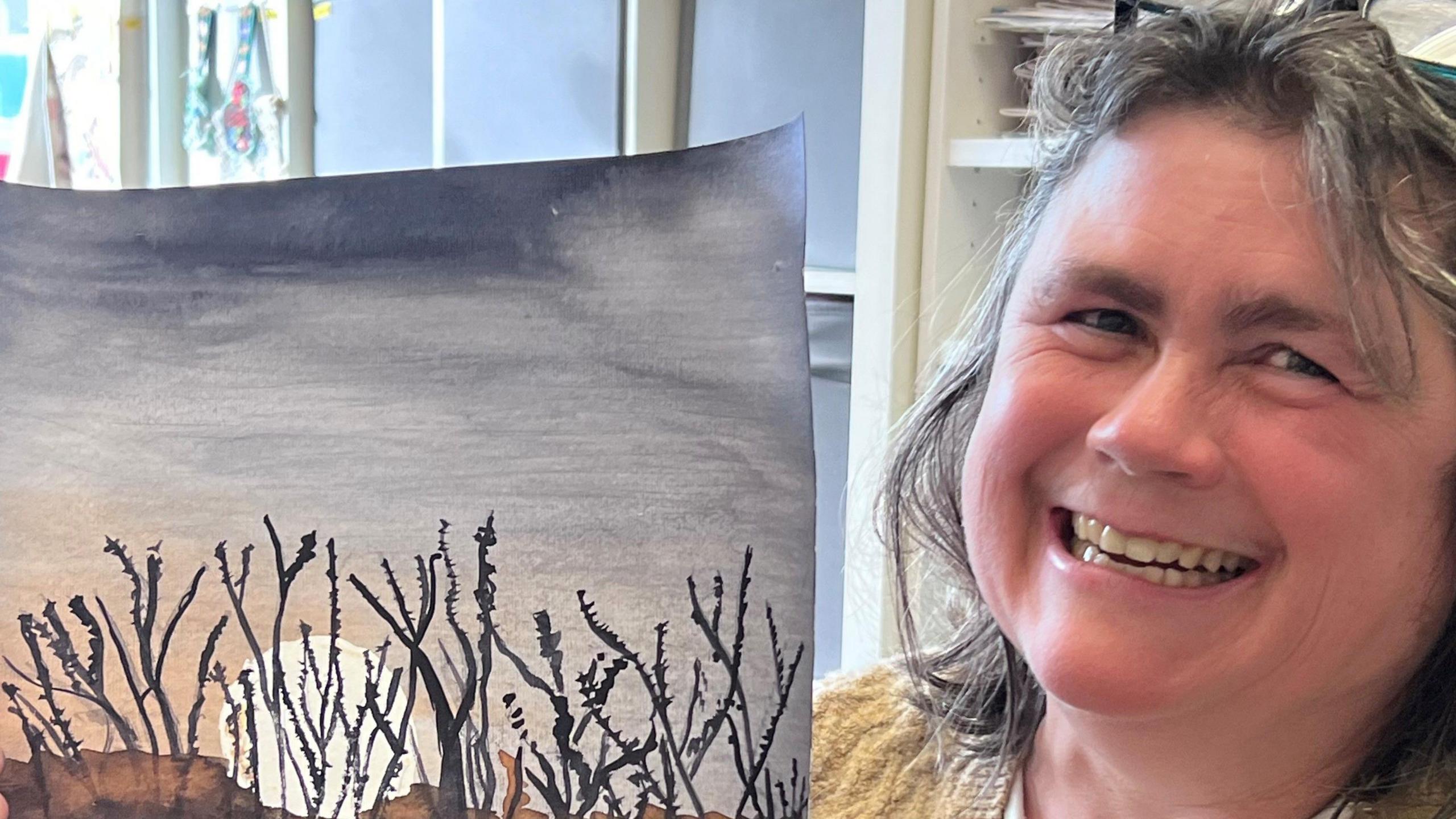 Manon has shoulder-length grey hair, is holding a painting of a sunset with vegetation in front of it and smiling at the camera.
