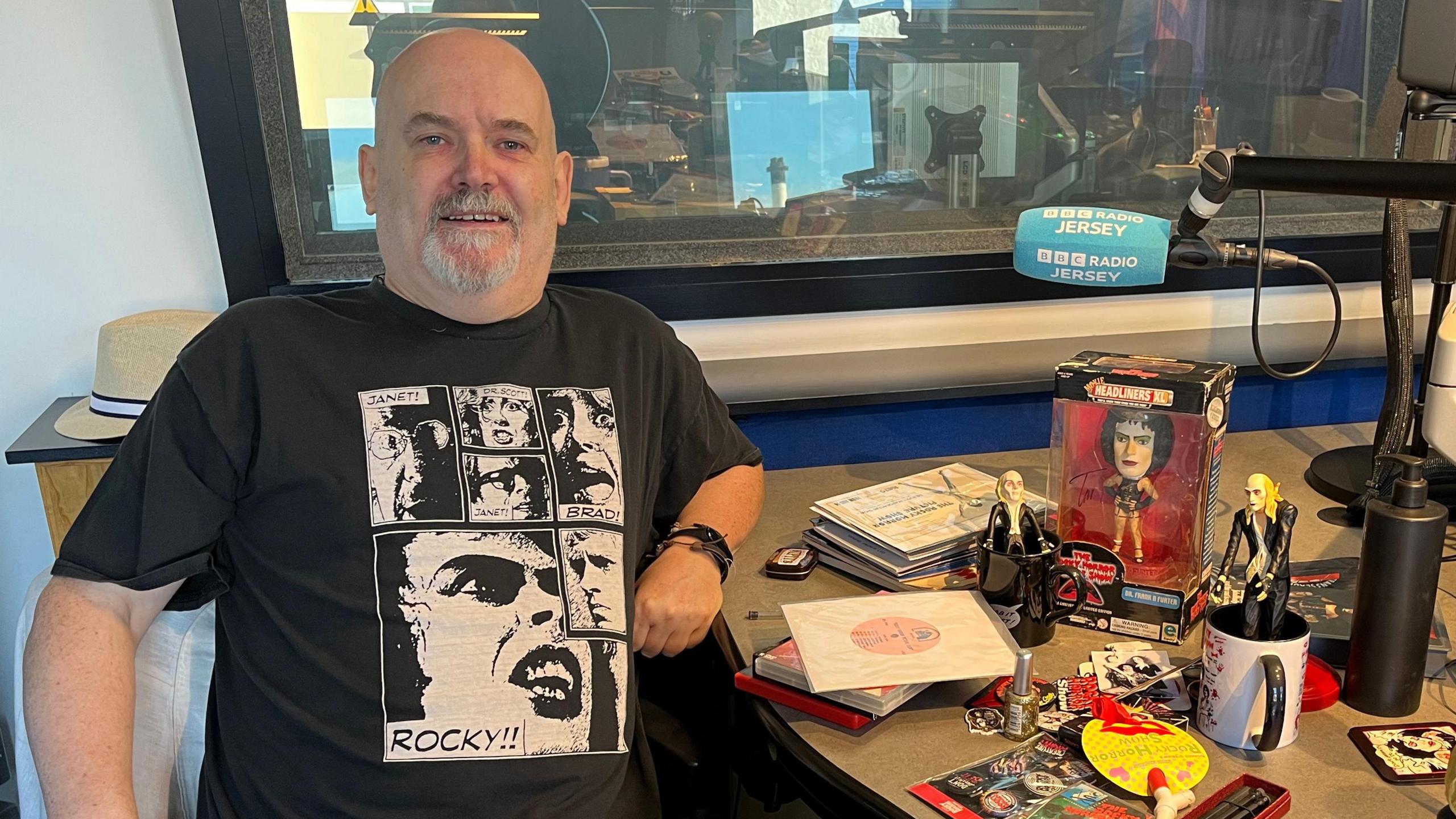 Glyn is seated in a radio studio, wearing a black 'Rocky!!' graphic T-shirt, surrounded by Rocky Horror memorabilia including figurines, books, and a mug on the table. Studio equipment and monitors are visible through a window behind them.